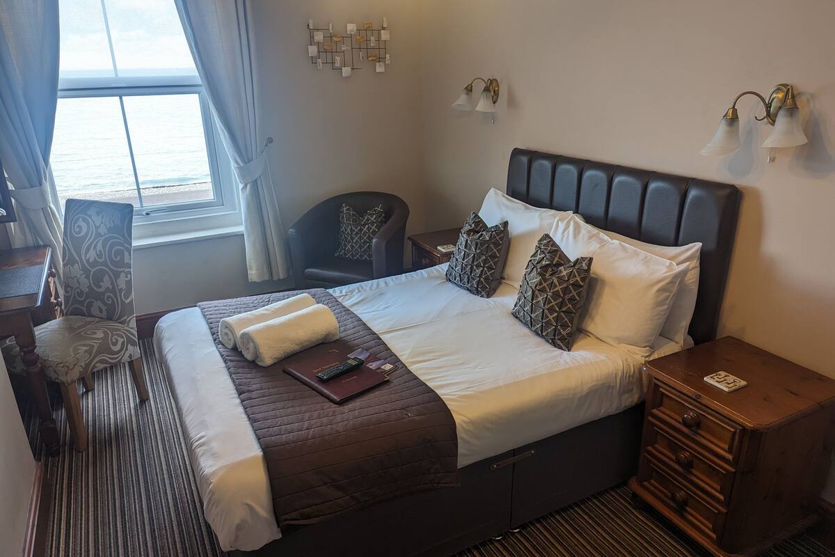 A neatly made bed with a brown headboard, a bedside table with a lamp, and a view of the sea through a window with curtains.