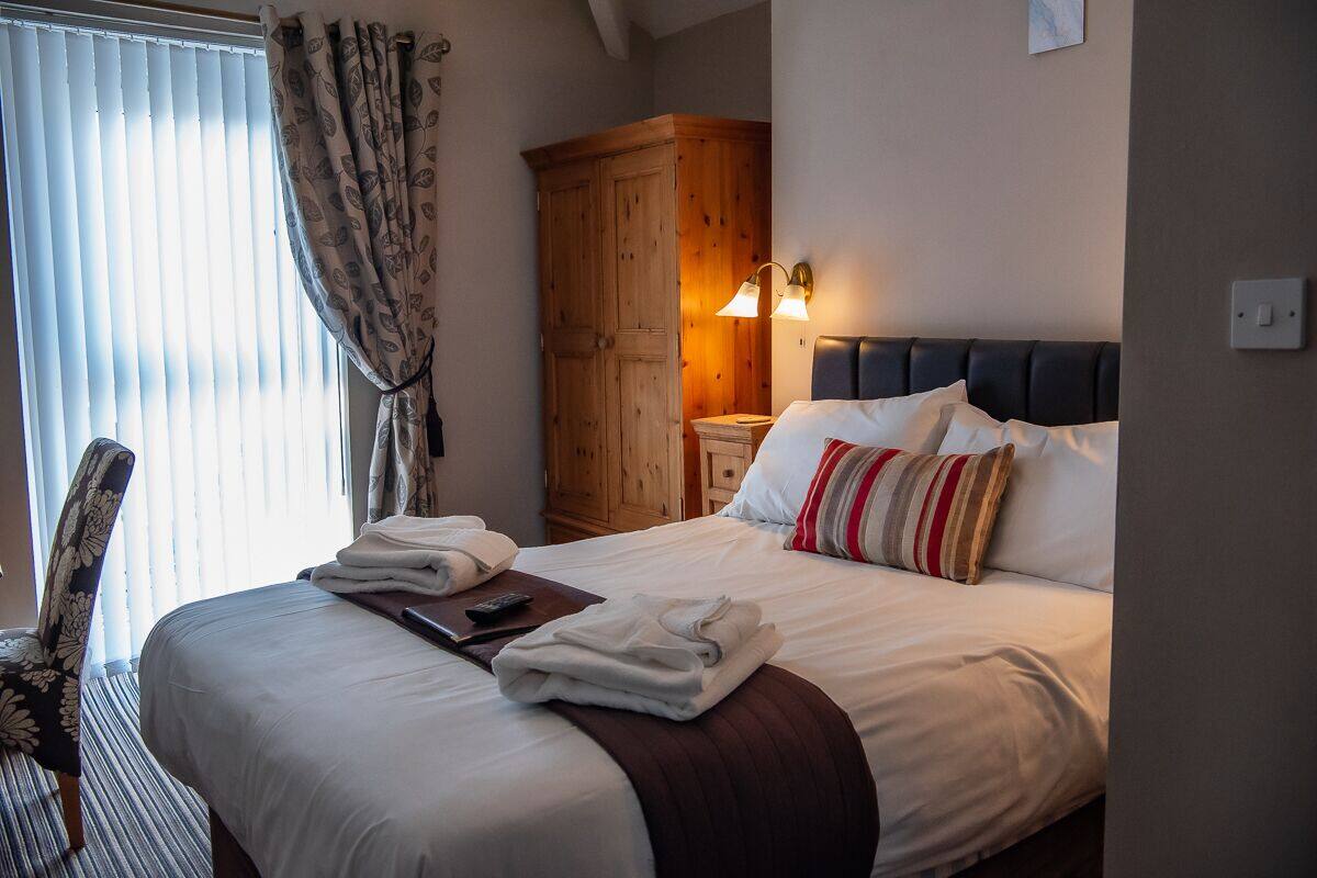 A hotel room with a neatly made bed, a wooden wardrobe, a nightstand with a lamp, and a chair with a floral pattern.