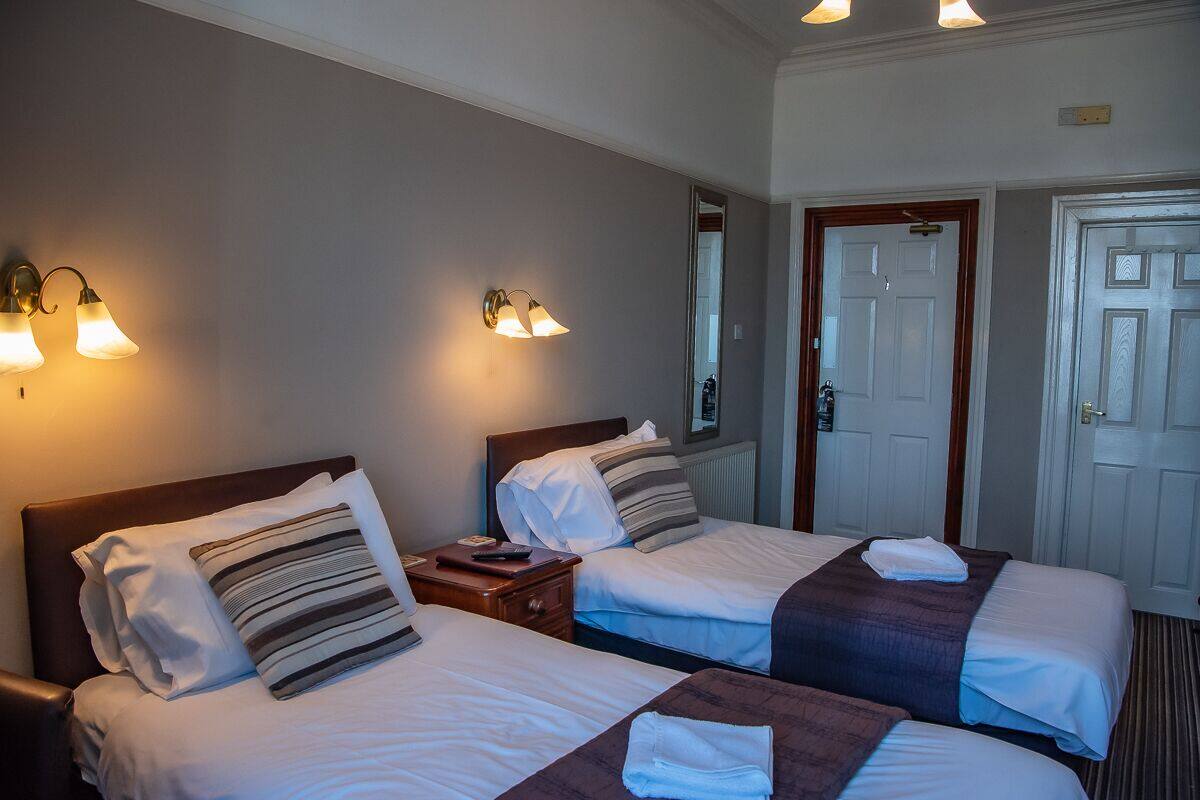 A hotel room with two beds, each with a pillow and a blanket, a nightstand with a lamp, and a door with a mirror.