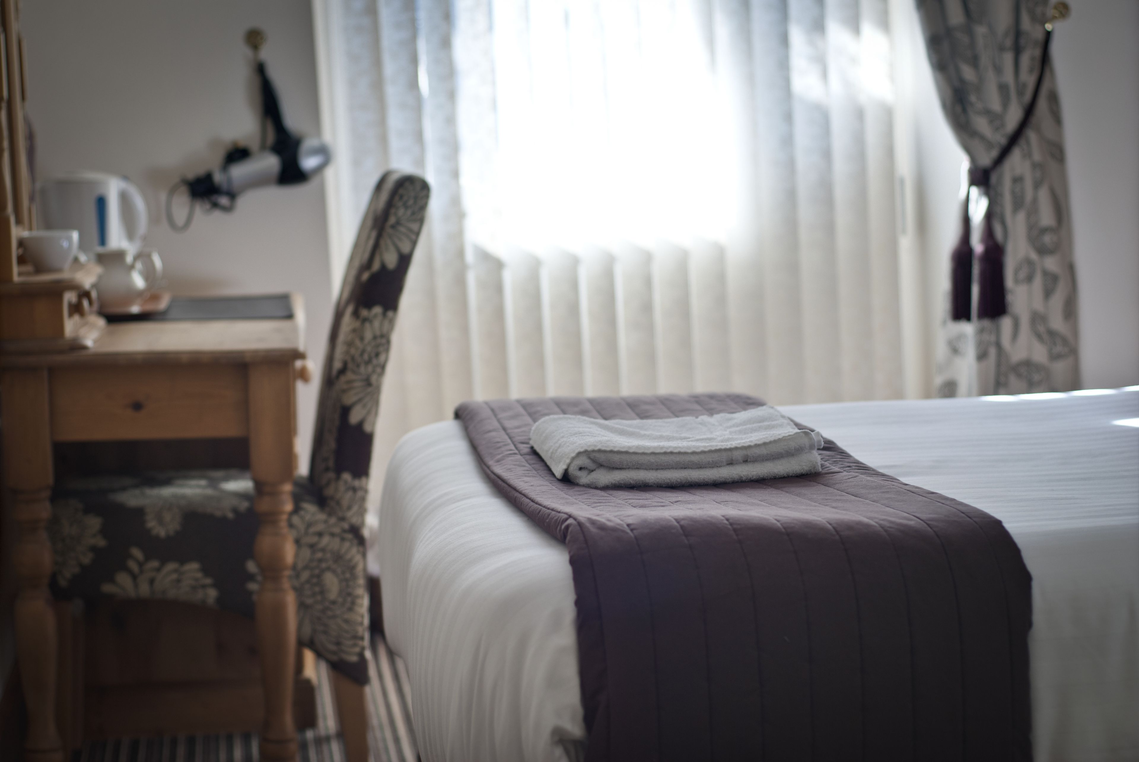 Un lit bien fait, avec une serviette pliée, une table de chevet en bois surmontée d’une lampe, et une chaise à motifs floraux.