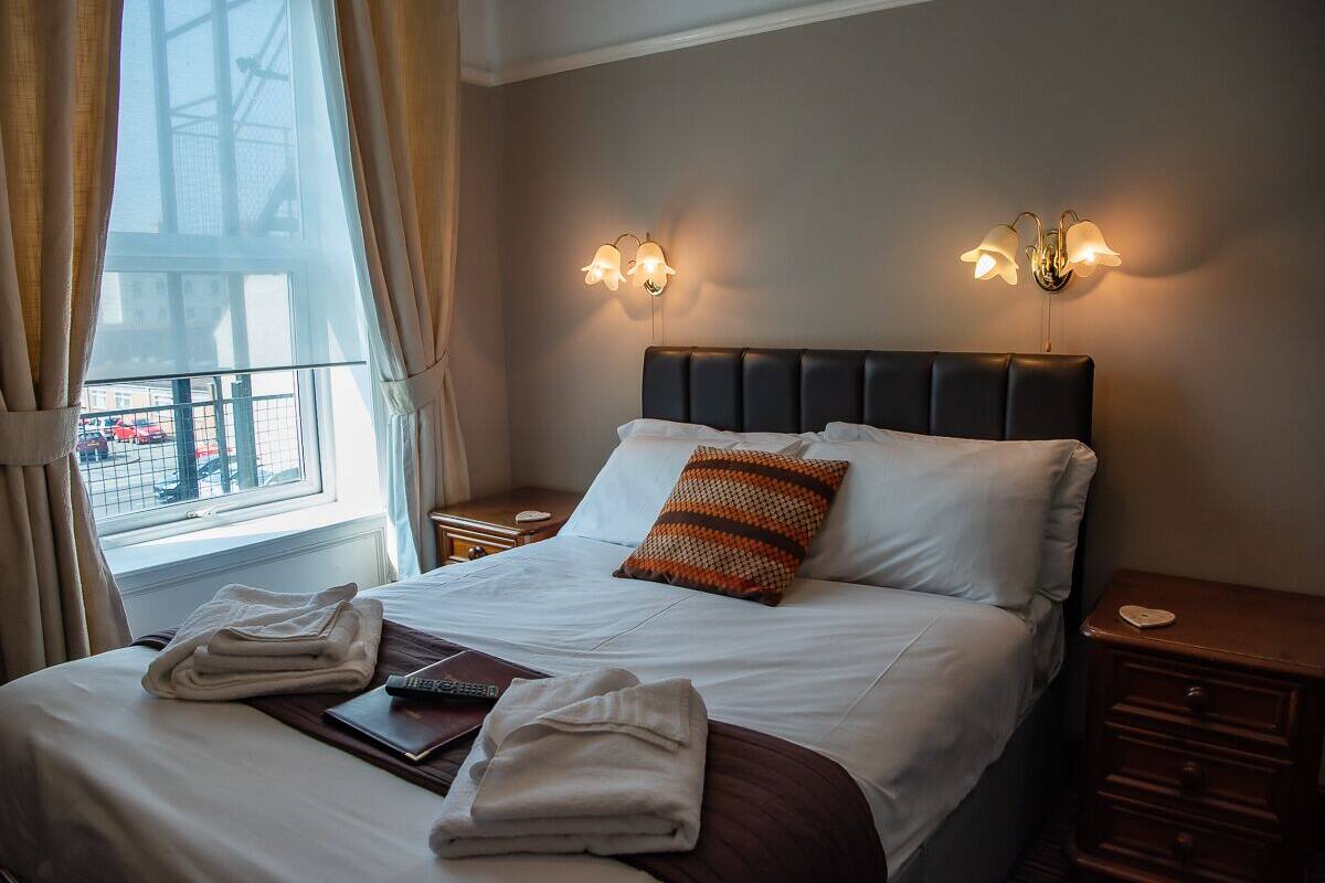 A neatly made bed with a striped pillow, two bedside lamps, a window with curtains, and a view of buildings outside.