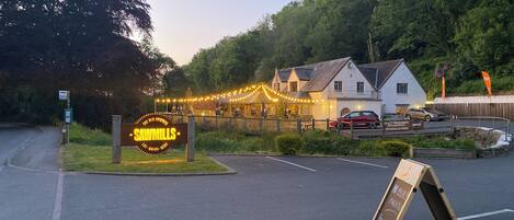 A sign for 'SAWMILLS' with a white building in the background, a parking area, and a grassy area with a signboard.