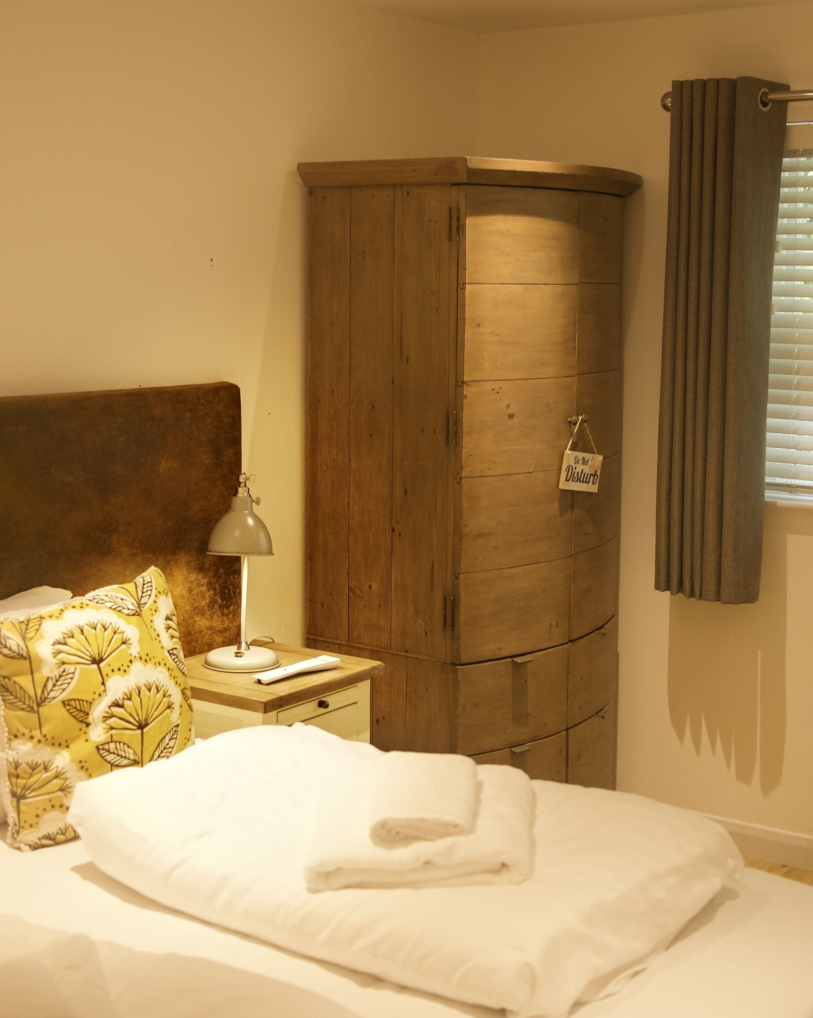 Une chambre avec une armoire en bois, une table de chevet avec une lampe, un lit avec du linge de maison blanc et un oreiller à motifs floraux.