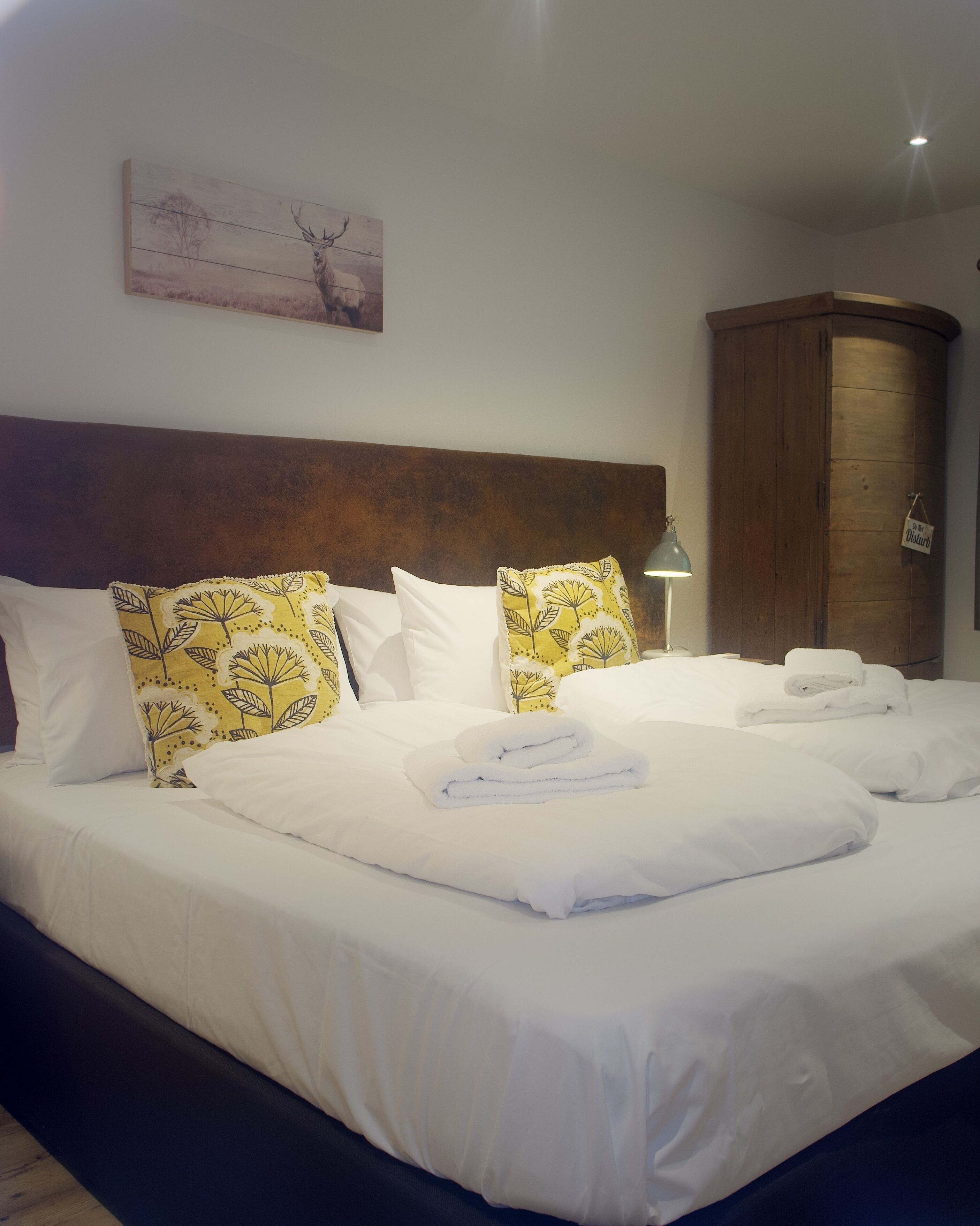 A neatly made bed with white linens and yellow patterned pillows, a wooden headboard, and a painting on the wall.