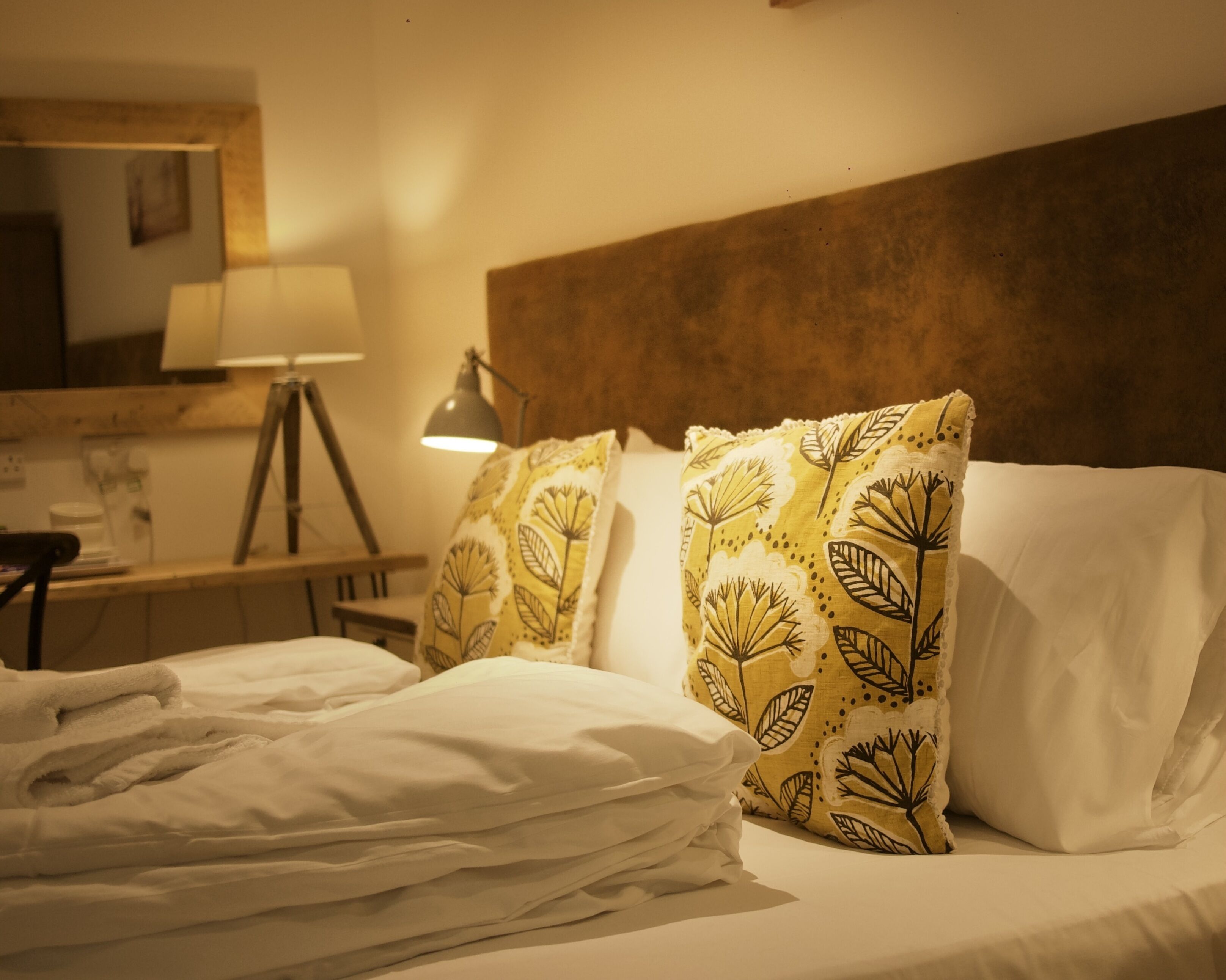 A neatly made bed with white linens, two decorative pillows with a leaf pattern, a bedside lamp, and a wooden headboard.