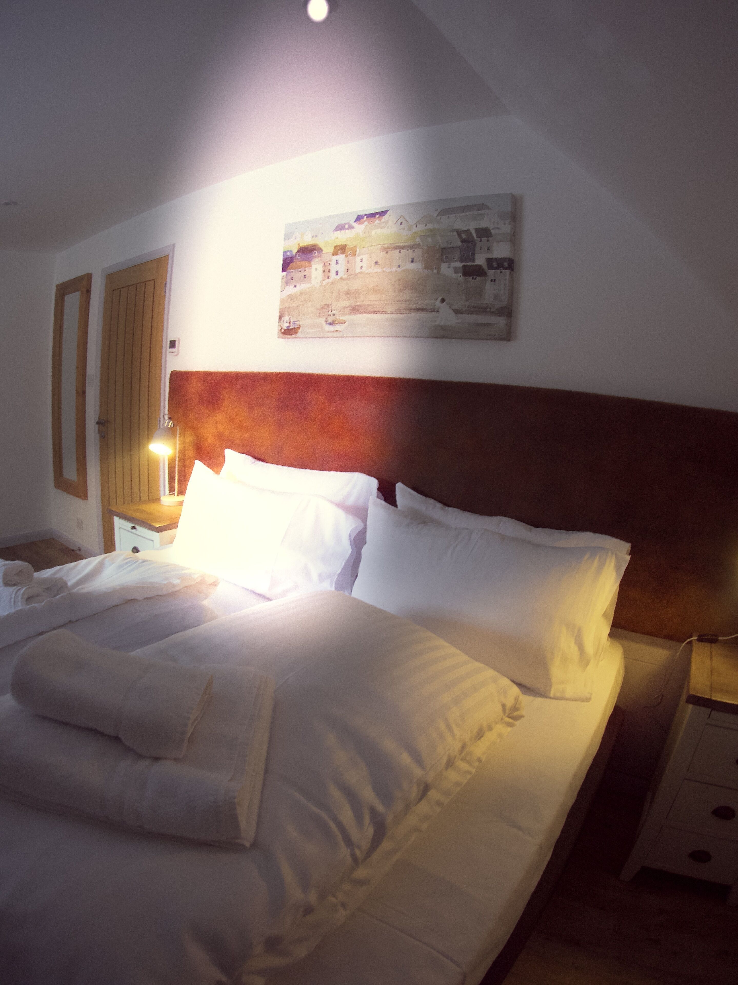 Un lit bien fait, avec du linge de lit blanc et des oreillers, une tête de lit en bois, un tableau accroché au mur et une lampe de chevet.