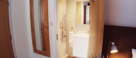 Un dormitorio con piso de madera, una cama con ropa de cama blanca, una mesita de noche con lámpara, y una puerta de madera que da a un baño con espejo, lavamanos y toallero.
