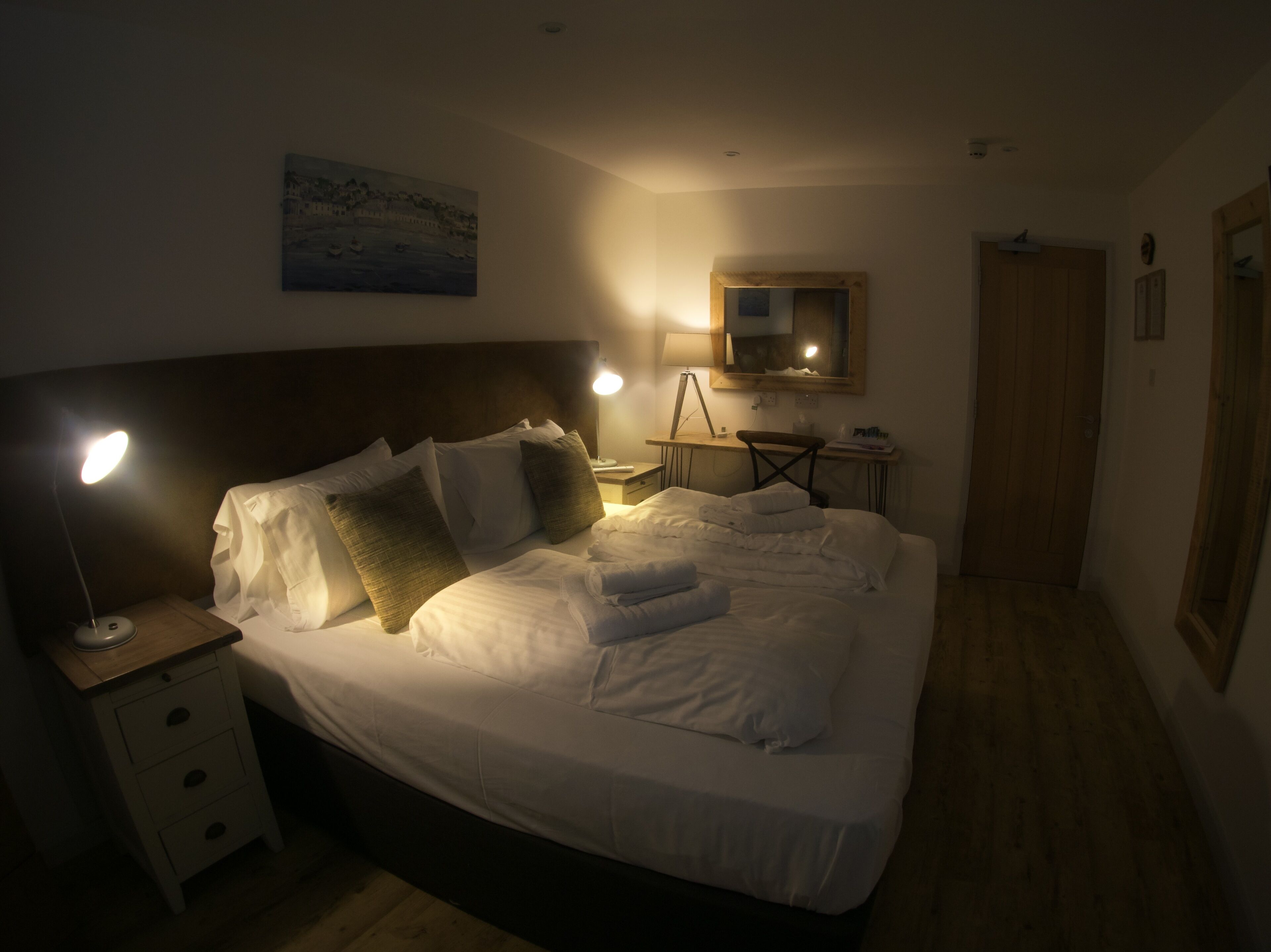 A neatly made bed with white linens and pillows, a bedside lamp, a small desk with a lamp, a chair, a wooden door, and a painting on the wall.