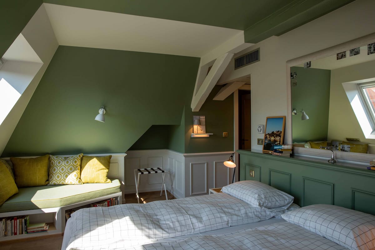 Un dormitorio con techo inclinado, una cama con colcha de cuadros blancos, pared verde y ventana que deja entrar luz natural.