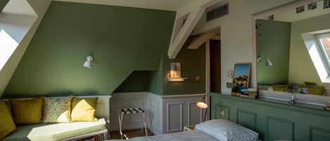 Un dormitorio con techo inclinado, una cama con colcha de cuadros blancos, pared verde y ventana que deja entrar luz natural.