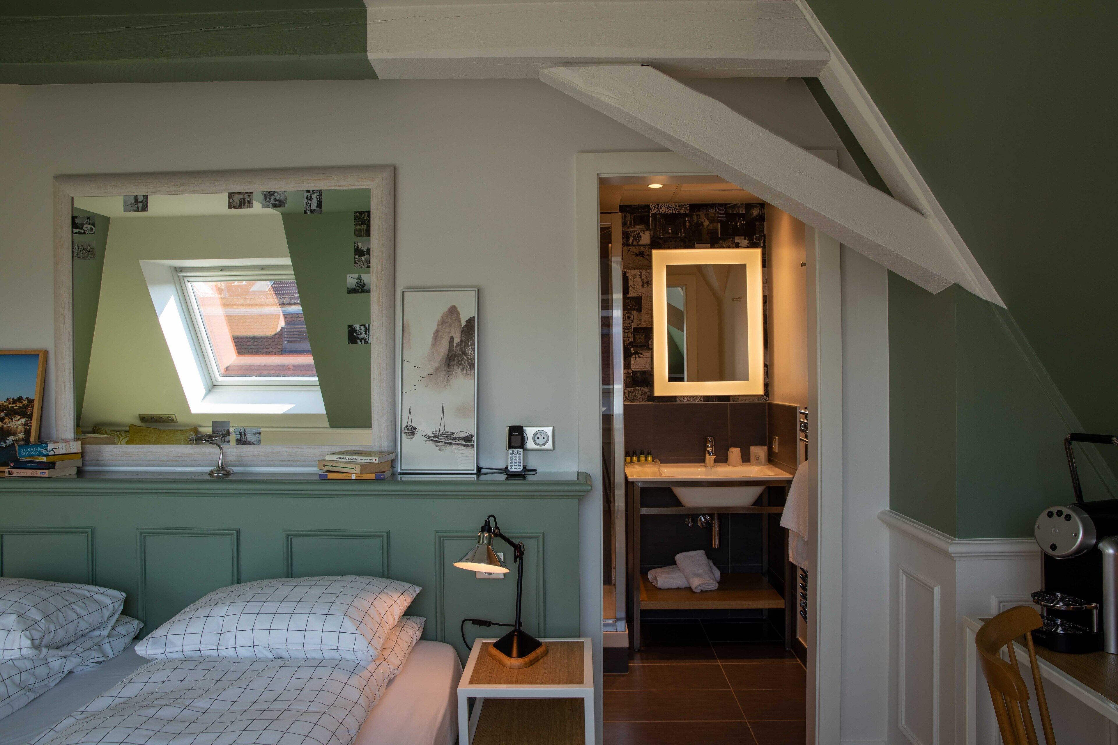 A bedroom with a skylight, a bedside table with a lamp, a bed with pillows, and a view of a bathroom with a mirror and sink.