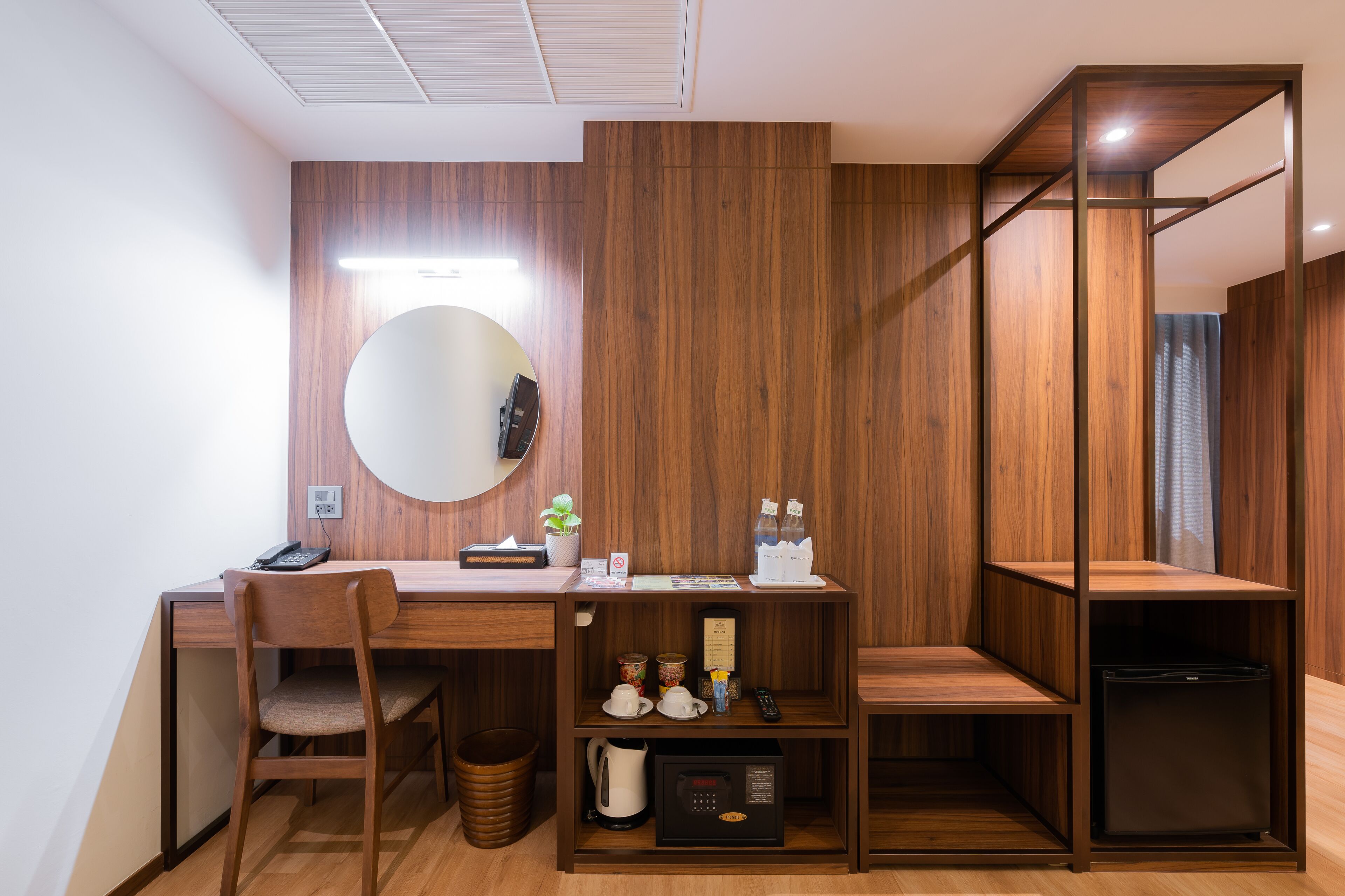 A compact hotel room with a wooden desk, a chair, a small refrigerator, a microwave, and a shelf with toiletries.