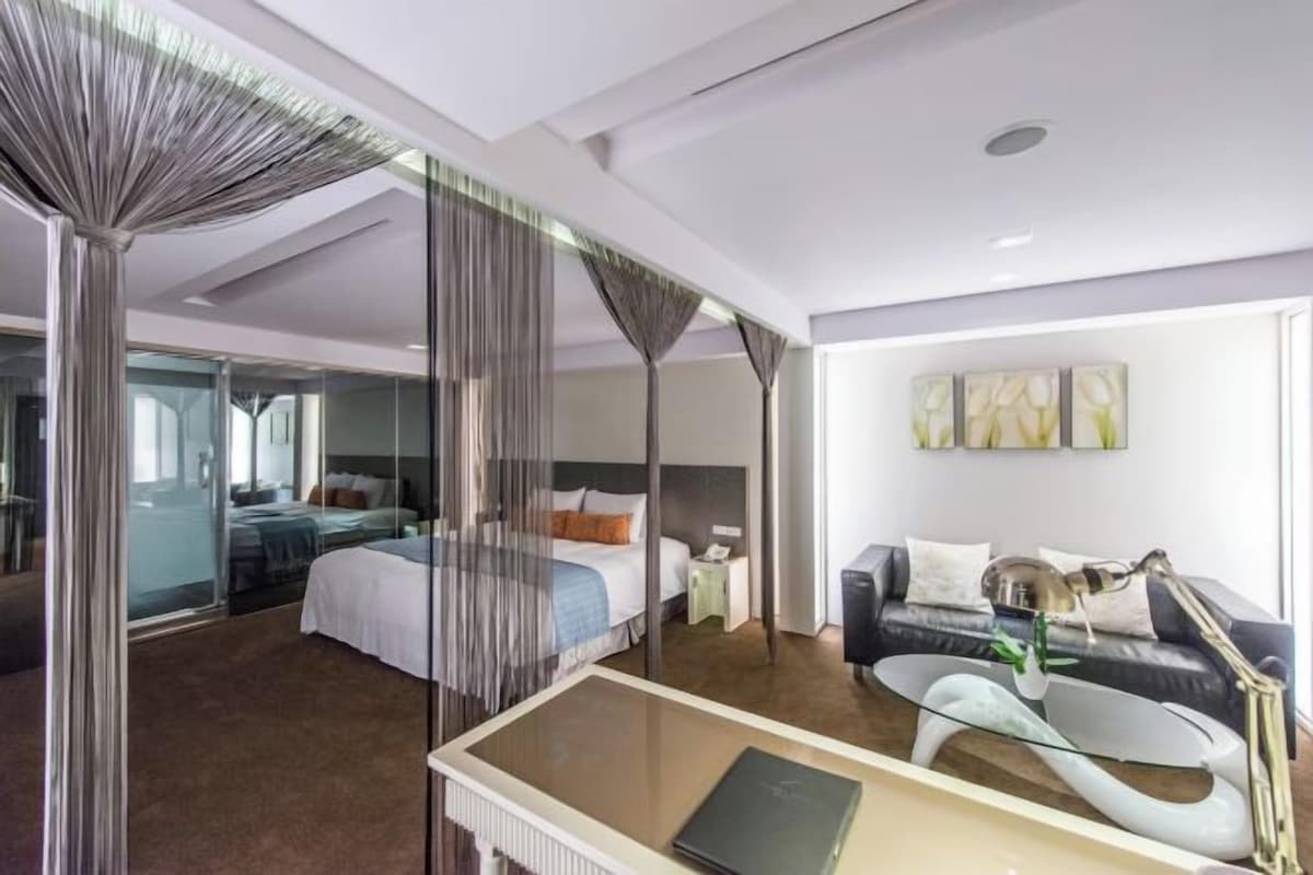 Habitación de hotel moderna con una cama grande, un sofá, un escritorio con lámpara y un separador de vidrio.