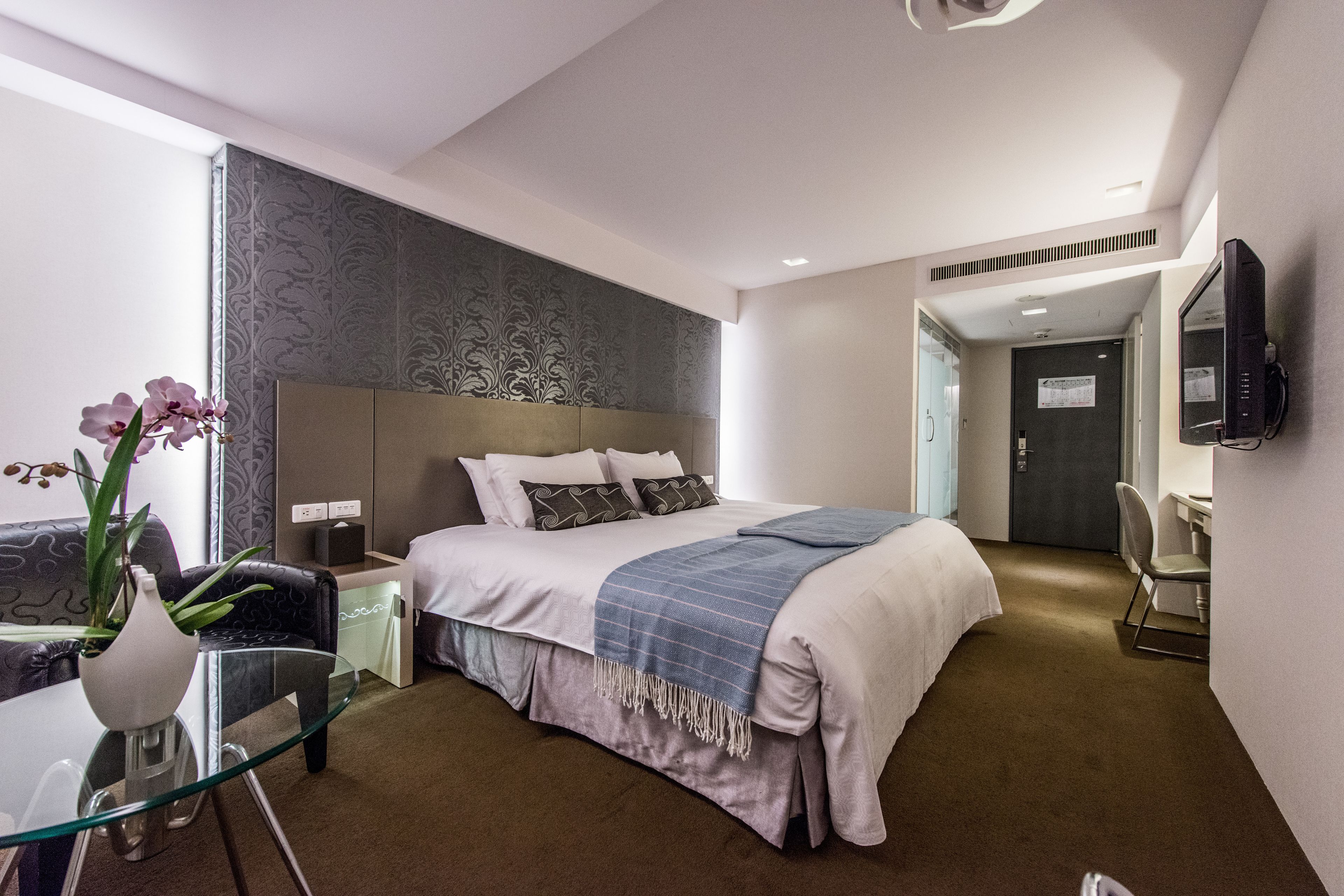 Una habitación de hotel moderna con una cama grande, una mesa de vidrio con un florero y un televisor montado en la pared, además de una puerta que da a otra habitación.