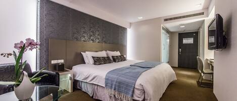 Una habitación de hotel moderna con una cama grande, una mesa de vidrio con un florero y un televisor montado en la pared, además de una puerta que da a otra habitación.