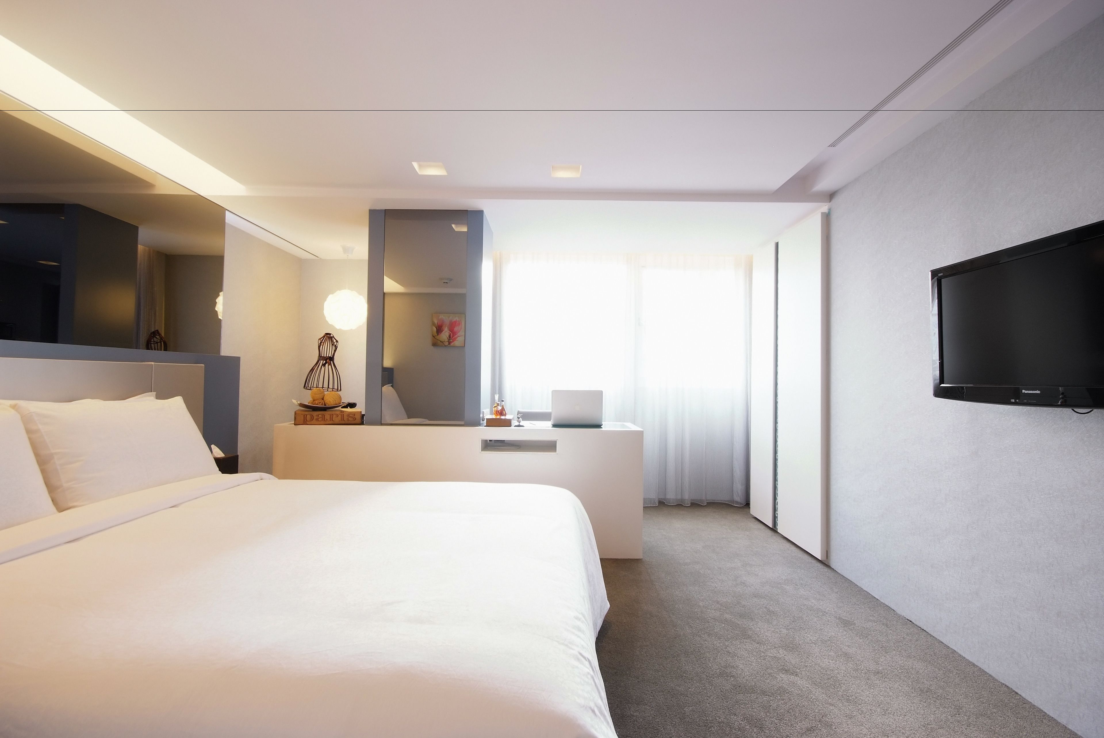 A modern hotel room with a large bed, a desk with a laptop, a flat-screen TV, and a large window with curtains.