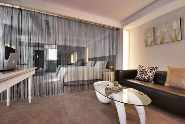 Una habitación de hotel moderna con una pared divisoria de vidrio, una computadora portátil sobre un escritorio, una mesa de centro de vidrio, un sofá de cuero negro y una cama con ropa blanca.
