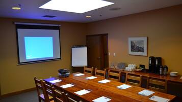 Un salón de conferencias con una larga mesa de madera, sillas, una pantalla de proyector, una pizarra y equipo para preparar café.
