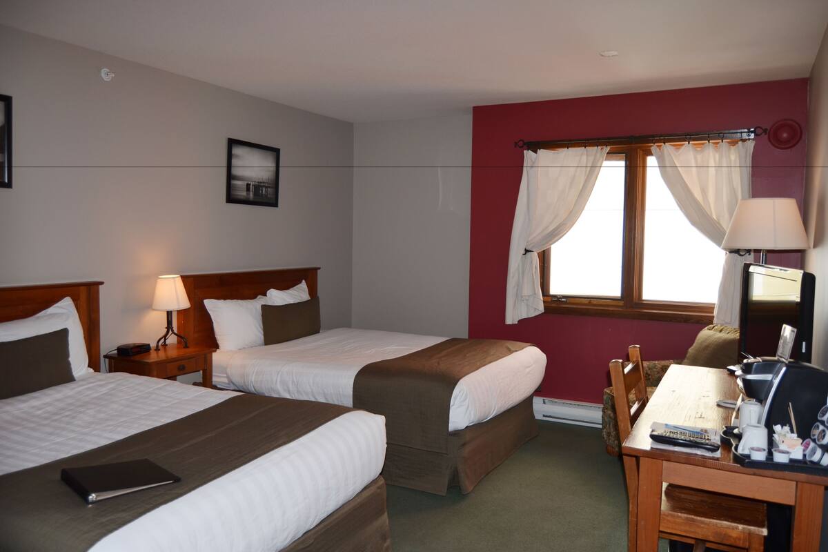 A hotel room with two beds, a desk with a chair, a lamp, and a window with curtains.