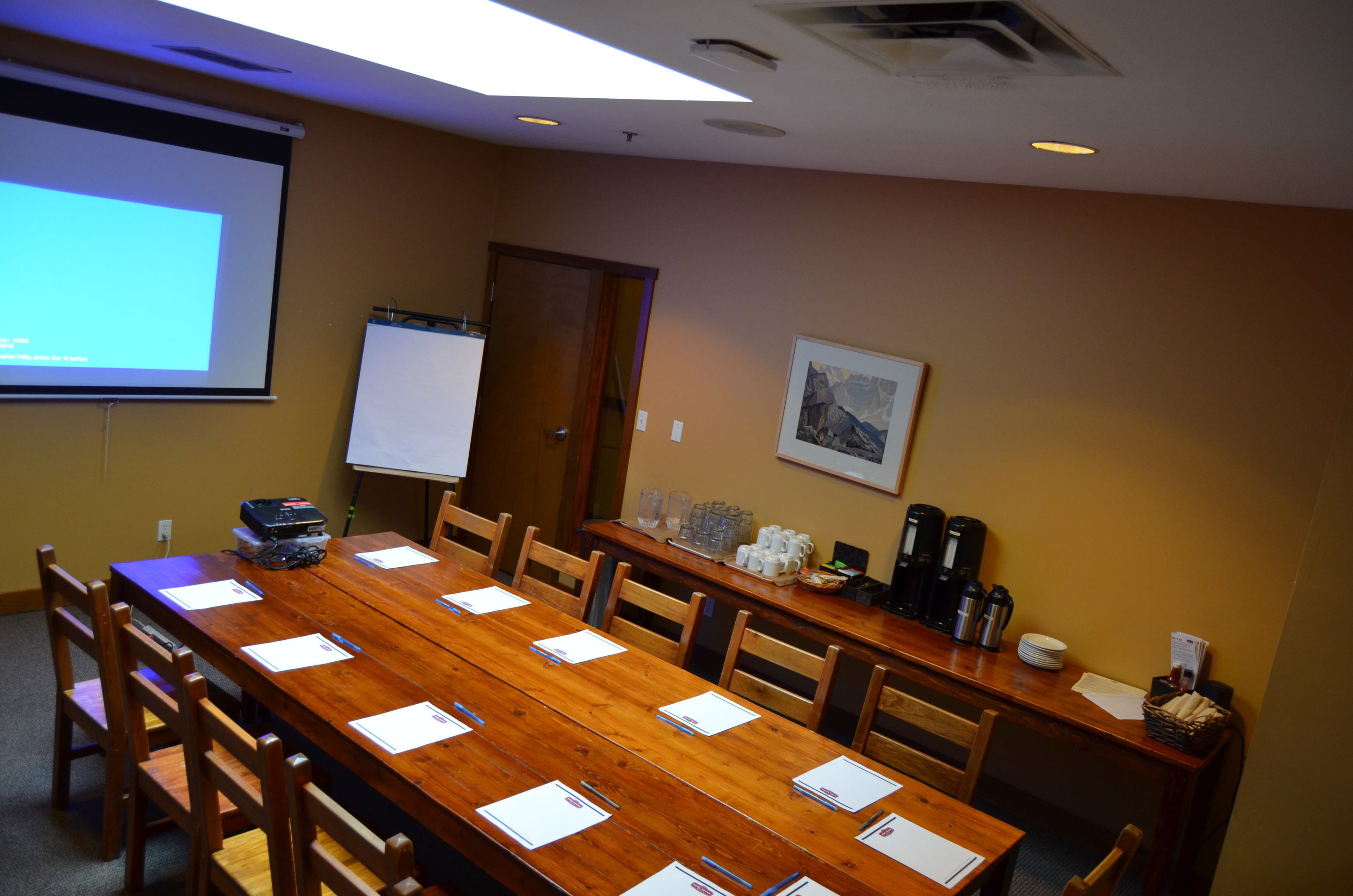 Un salón de conferencias con una larga mesa de madera, sillas, una pantalla de proyector, una pizarra y una estación de café.