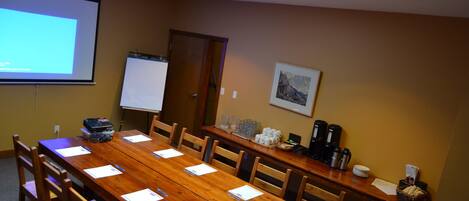 Un salón de conferencias con una larga mesa de madera, sillas, una pantalla de proyector, una pizarra y una estación de café.