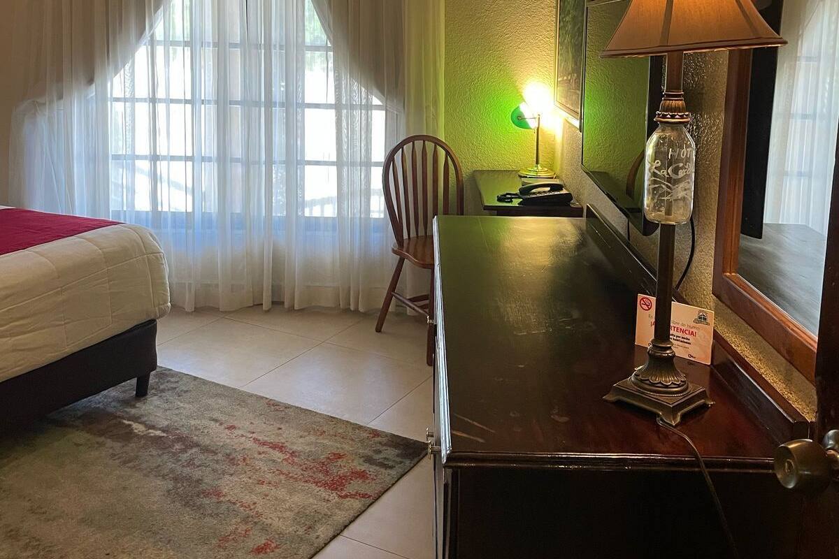 A hotel room with a bed, a desk, a chair, a lamp, and a large window with curtains.