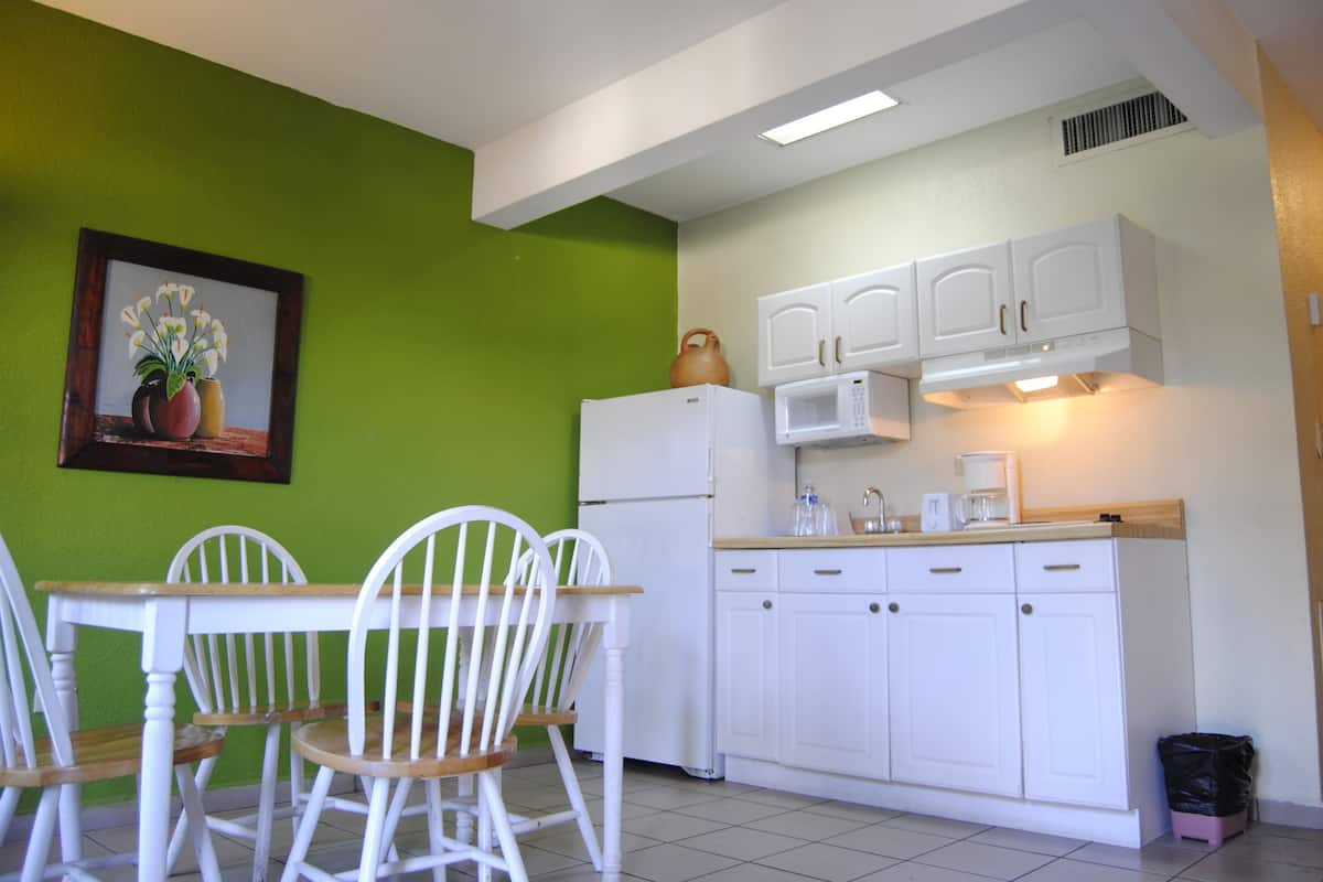 A compact kitchen with white cabinets, a refrigerator, a microwave, and a sink. There is a dining table with chairs and a framed picture on the wall.