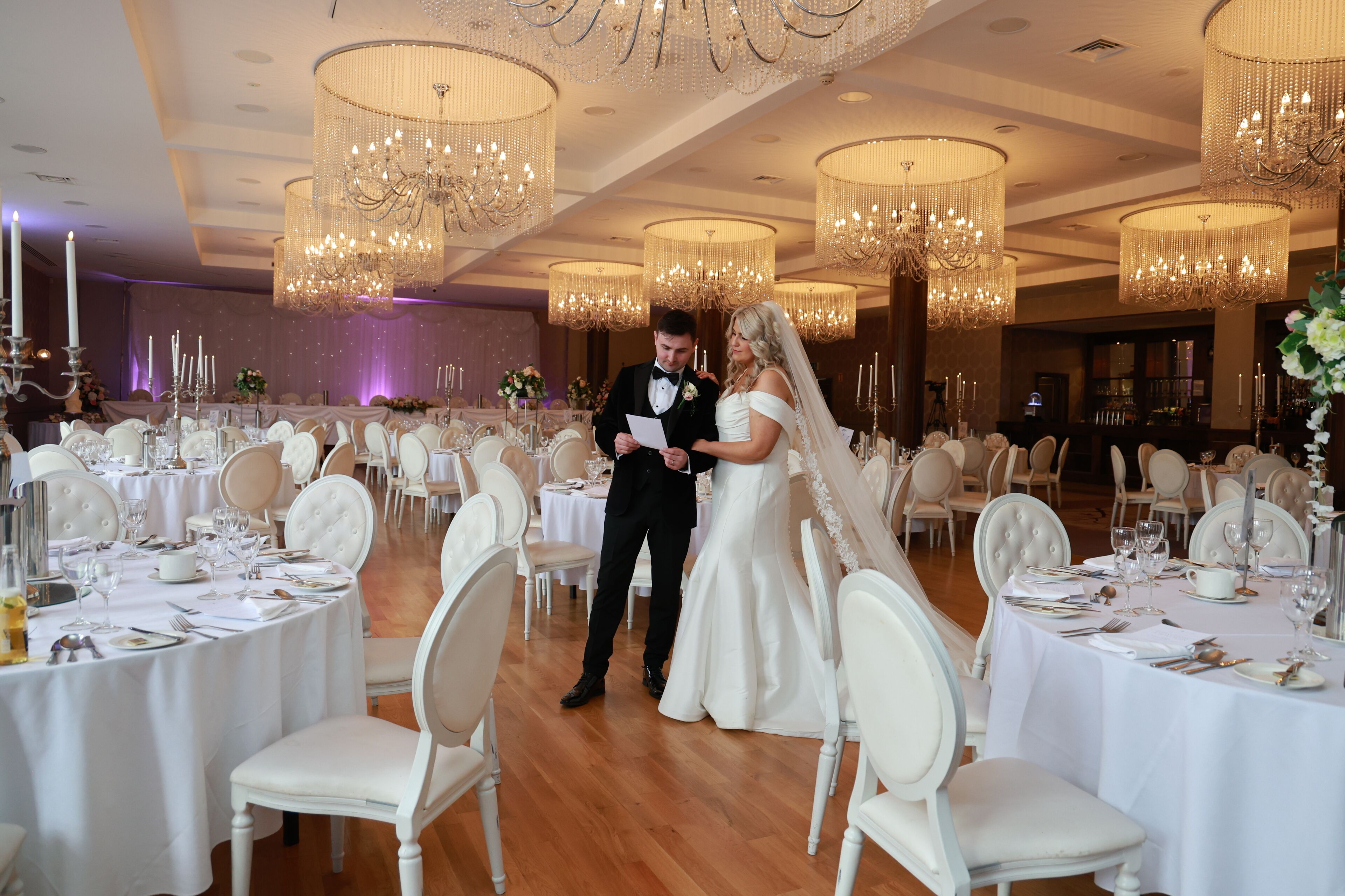 A wedding reception with round tables, white chairs, and chandeliers.