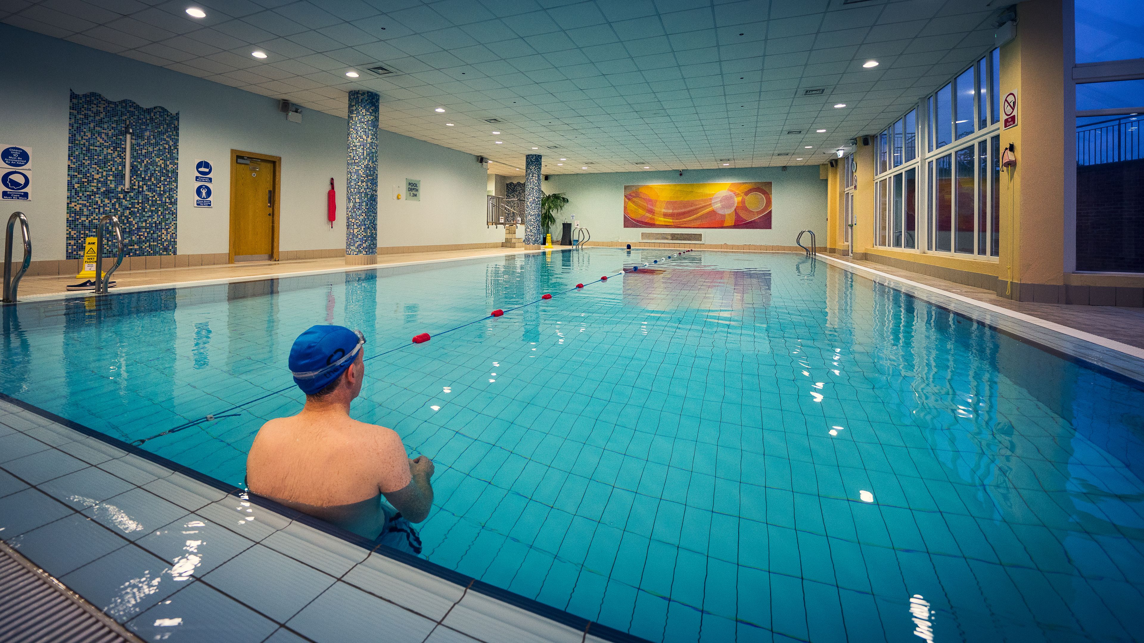 En indendørs swimmingpool med fliset gulv, en trædør og et farverigt muralmaleri på væggen.