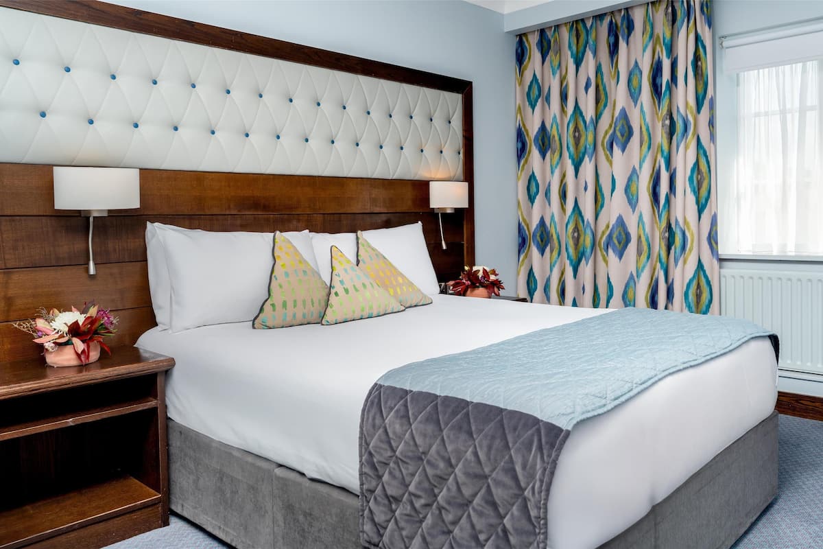 A hotel room with a large bed, a wooden headboard, a bedside table with a lamp, and patterned curtains.