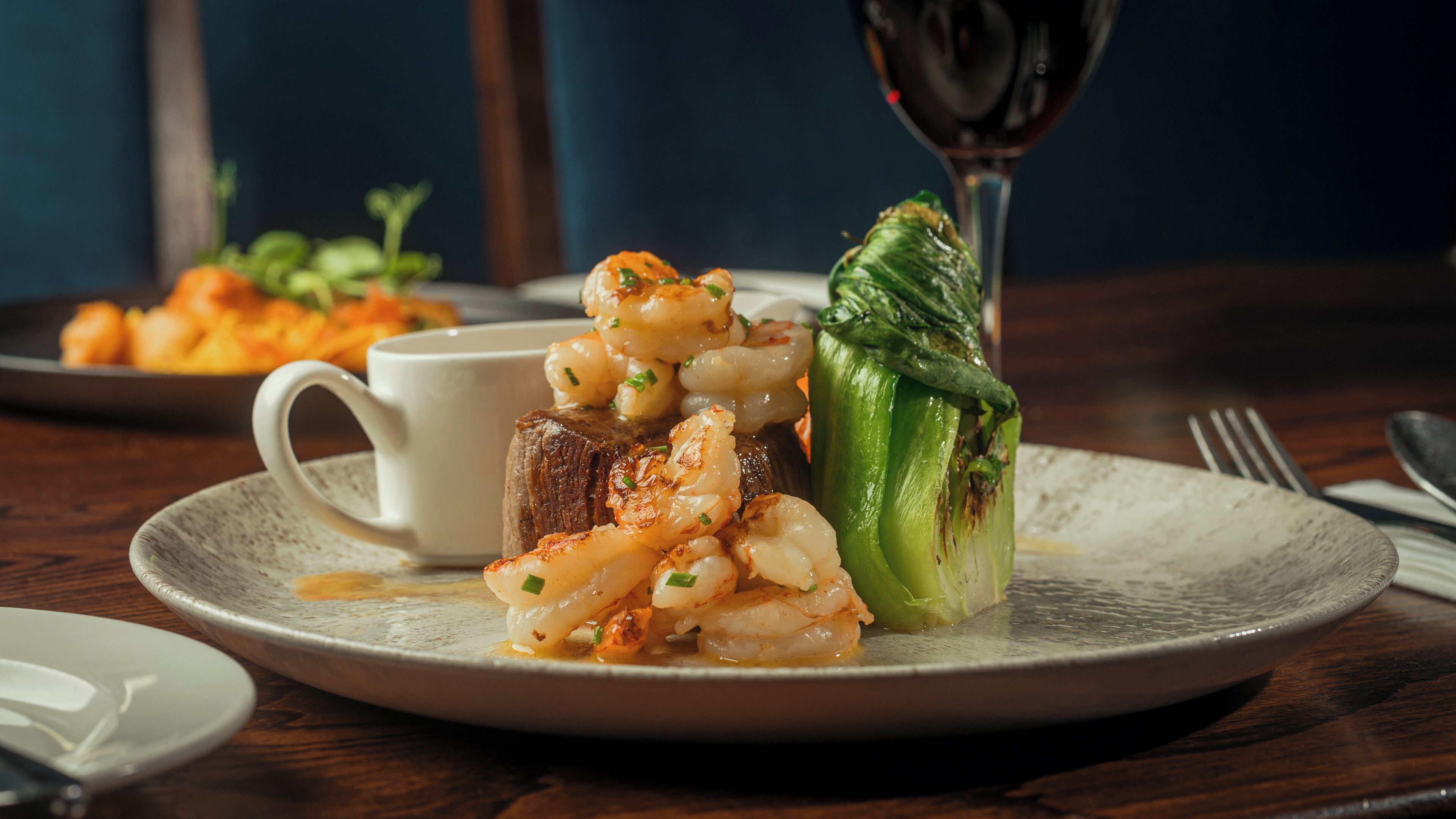 A plate with grilled shrimp, a piece of meat, and a stuffed vegetable, accompanied by a glass of red wine.