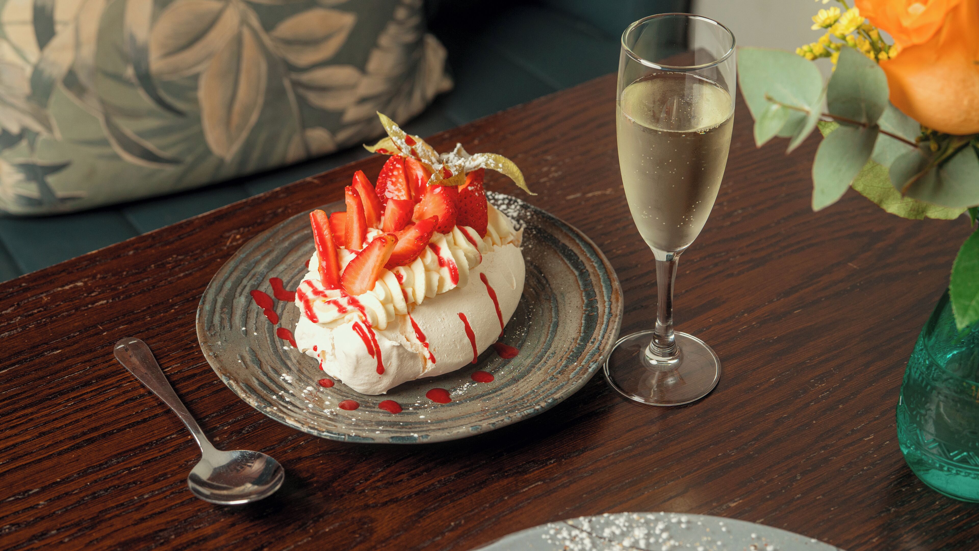 A dessert on a plate with a spoon beside it, a glass of a clear liquid, and a vase with flowers.