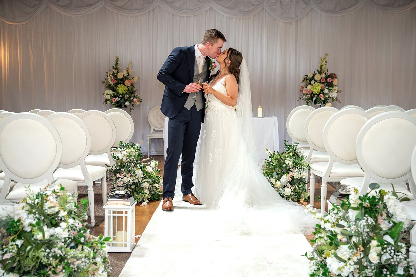 Une cérémonie de mariage où les mariés s’embrassent, entourés de chaises blanches et de compositions florales.