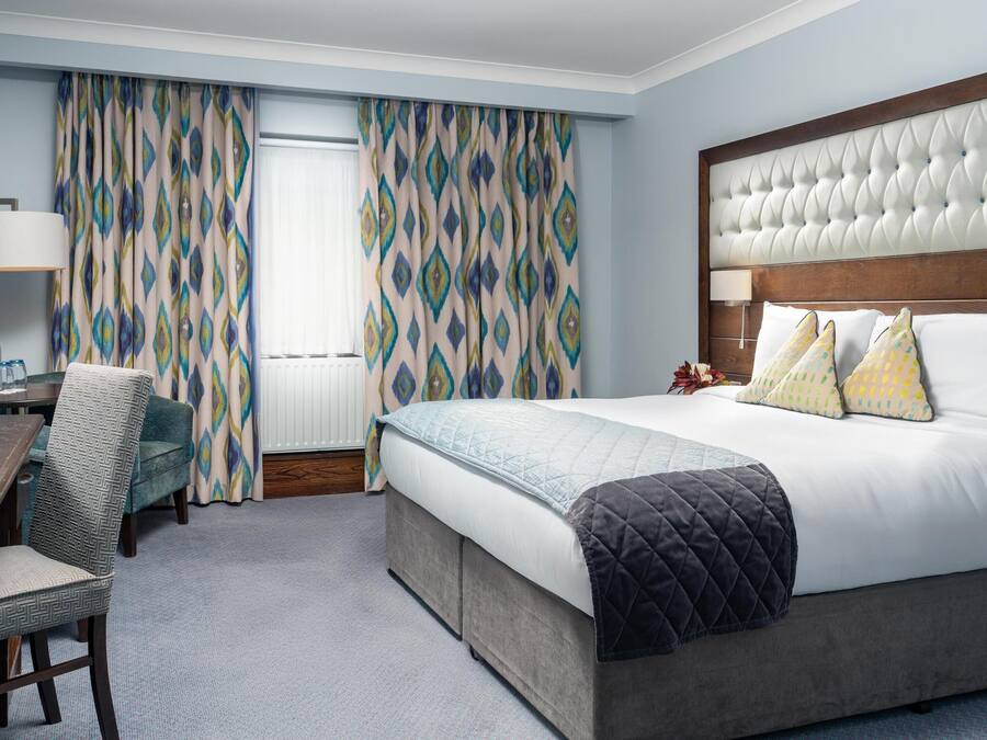 Habitación de hotel con una cama grande, cabecero de madera, escritorio con silla y cortinas estampadas.