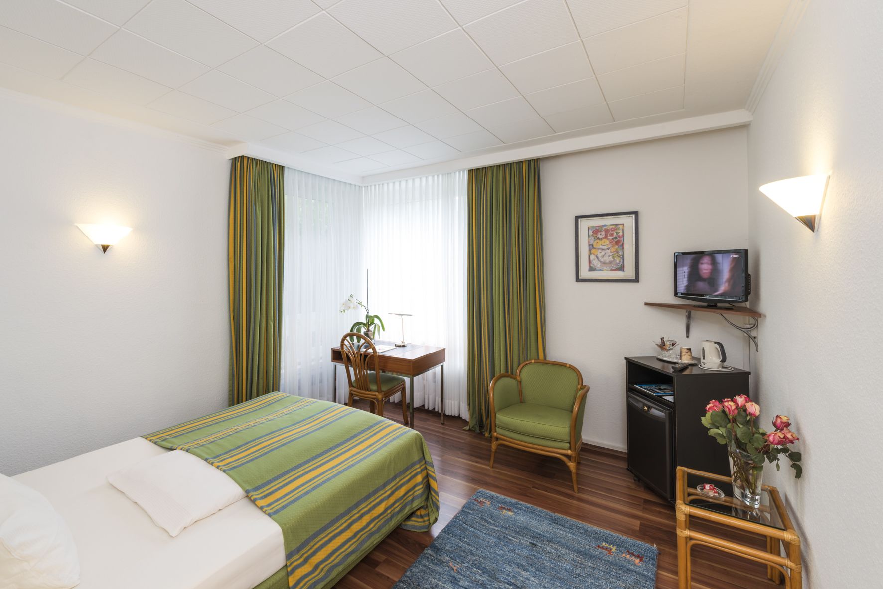 Ein Hotelzimmer mit einem Bett, einem Schreibtisch, einem Stuhl, einem Fernseher und einem kleinen Tisch mit Blumen.