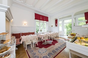 Uma sala de jantar com esquema de cores vermelho e branco, apresentando uma mesa comprida com cadeiras brancas, um tapete estampado e um buffet com diversos alimentos.