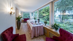 A dining area with a long table set for a meal, large windows with a view of greenery, and red upholstered chairs. - Parkhotel Atlantic Heidelberg (Heidelberg)