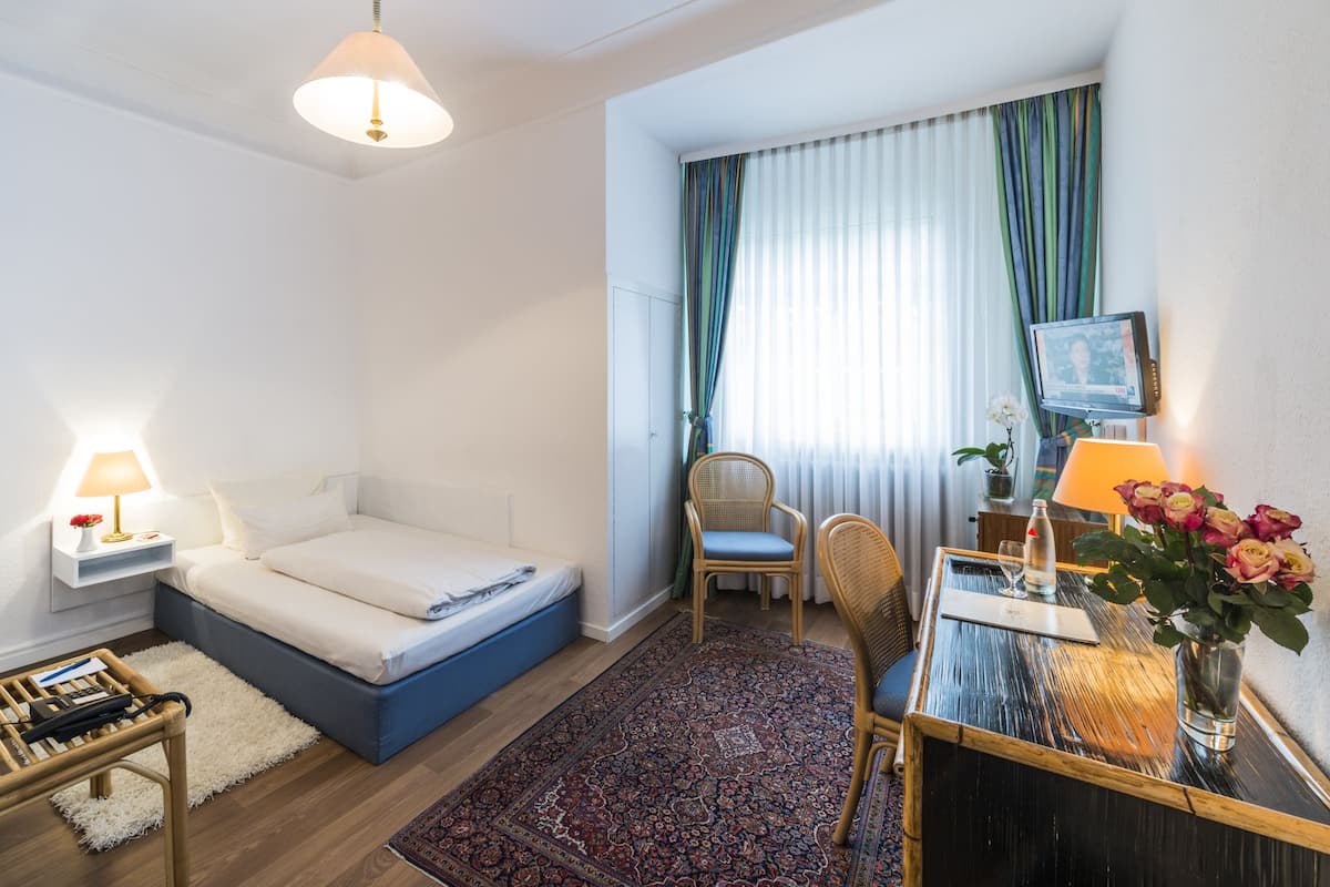 A hotel room with a bed, a desk with a lamp, a chair, a TV, and a window with curtains.