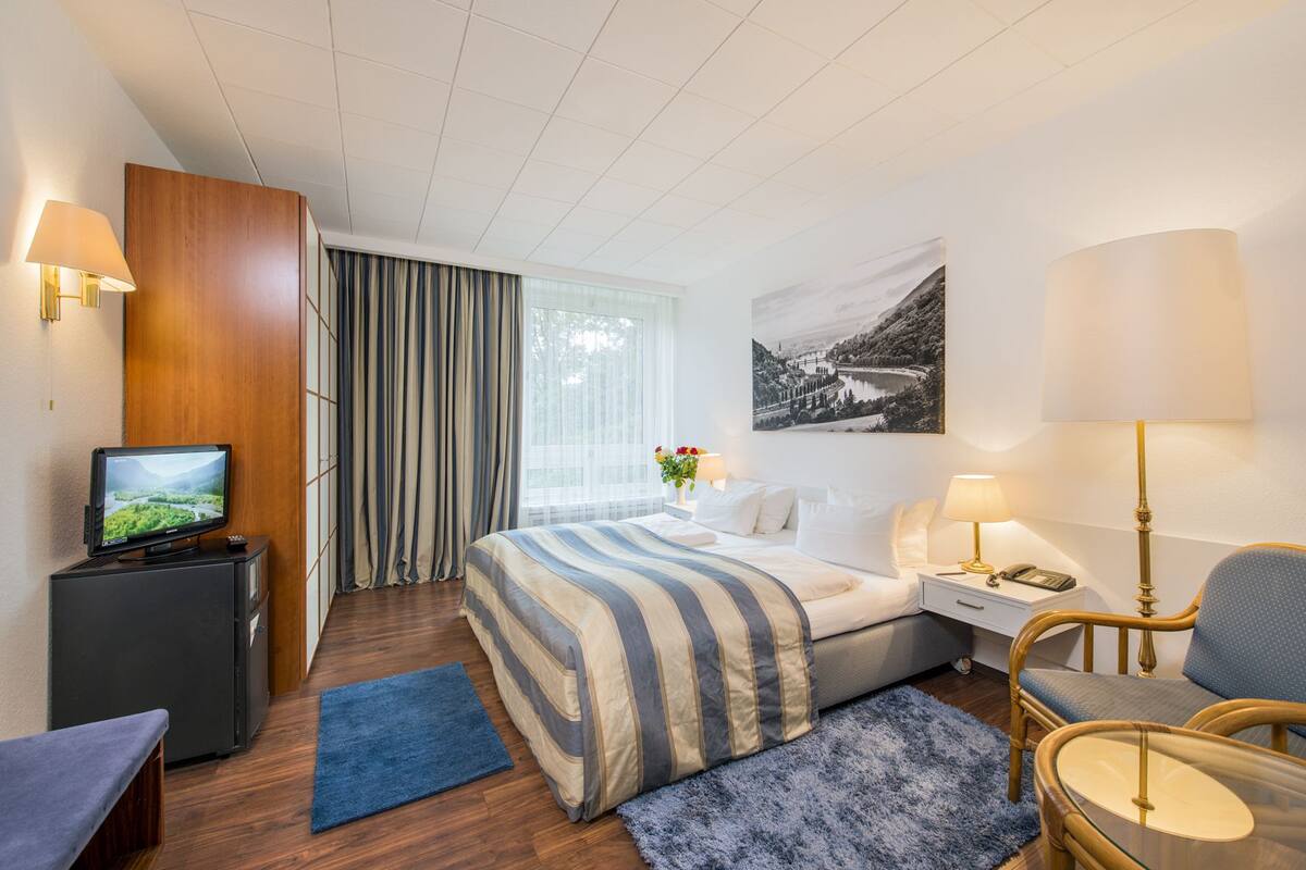 A hotel room with a bed, a desk, a chair, a TV, and a window with curtains.