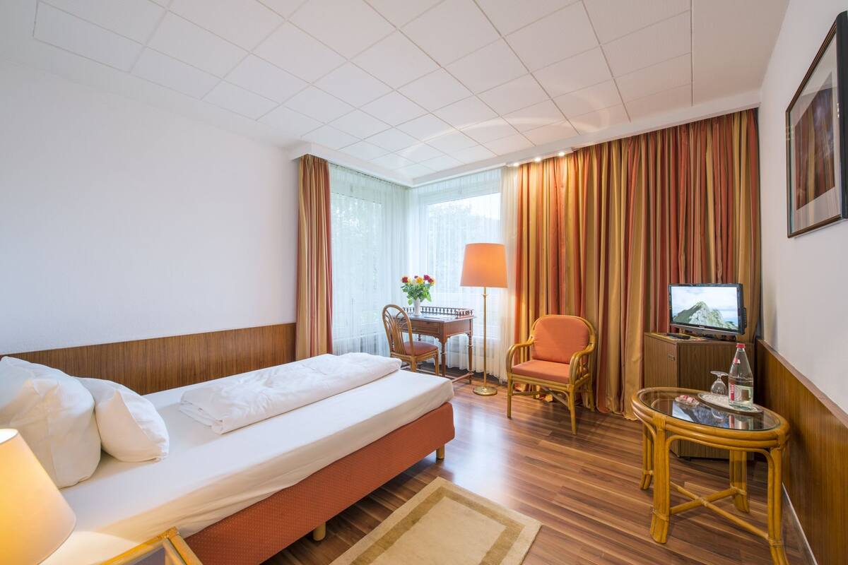 A hotel room with a single bed, a desk with a chair, a television, and a small table with a lamp.