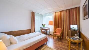 A hotel room with a single bed, a desk with a chair, a television, and a small table with a lamp.