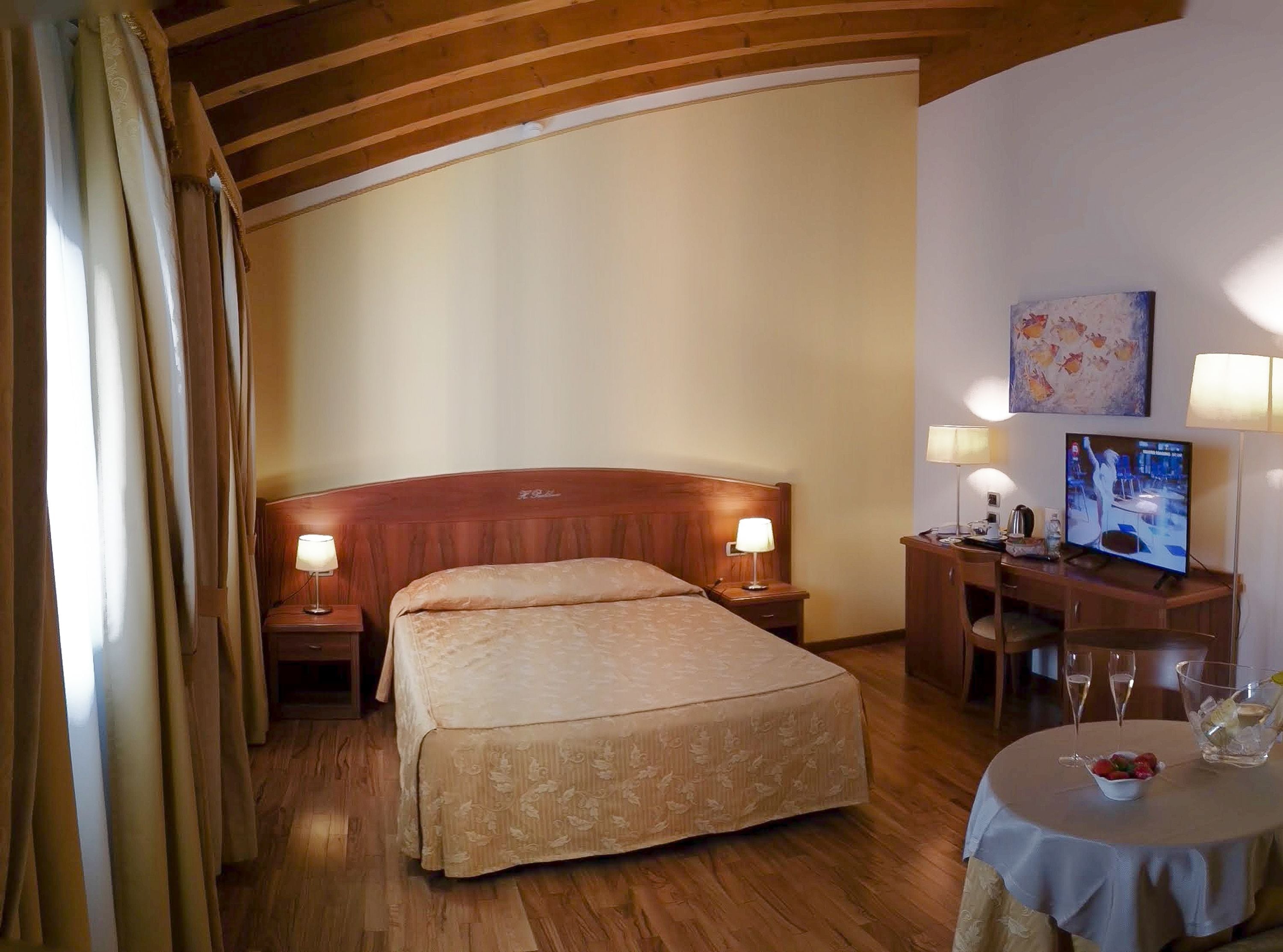 A hotel room with a wooden bed, two bedside lamps, a desk with a computer, a TV, and a small table with glasses and a bowl of fruit.