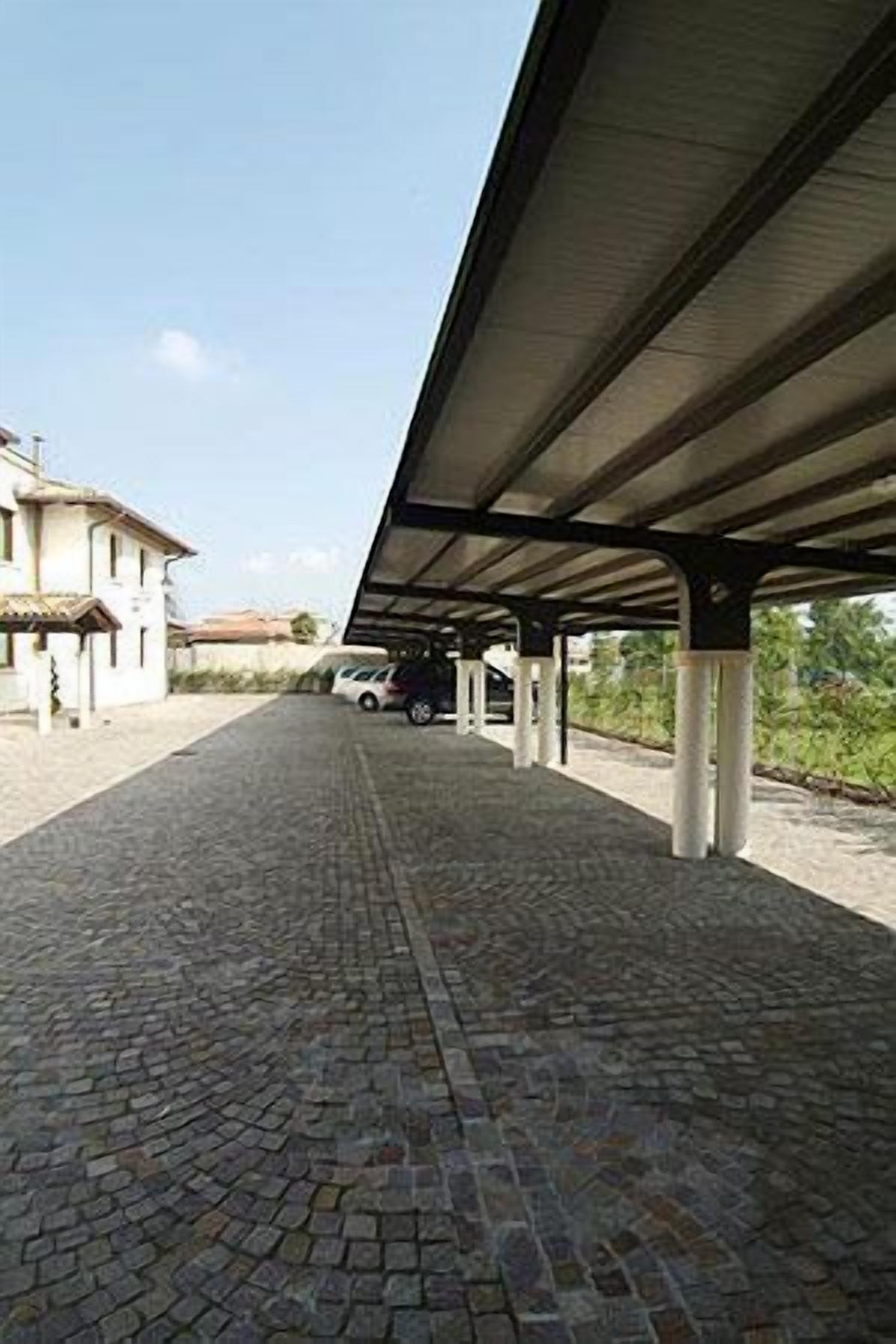 A covered outdoor area with a cobblestone floor, a car parked under a shelter, and a building in the background.