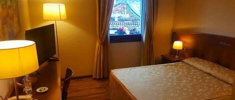 A hotel room with a bed, a desk with a lamp, a TV, and a window with curtains.