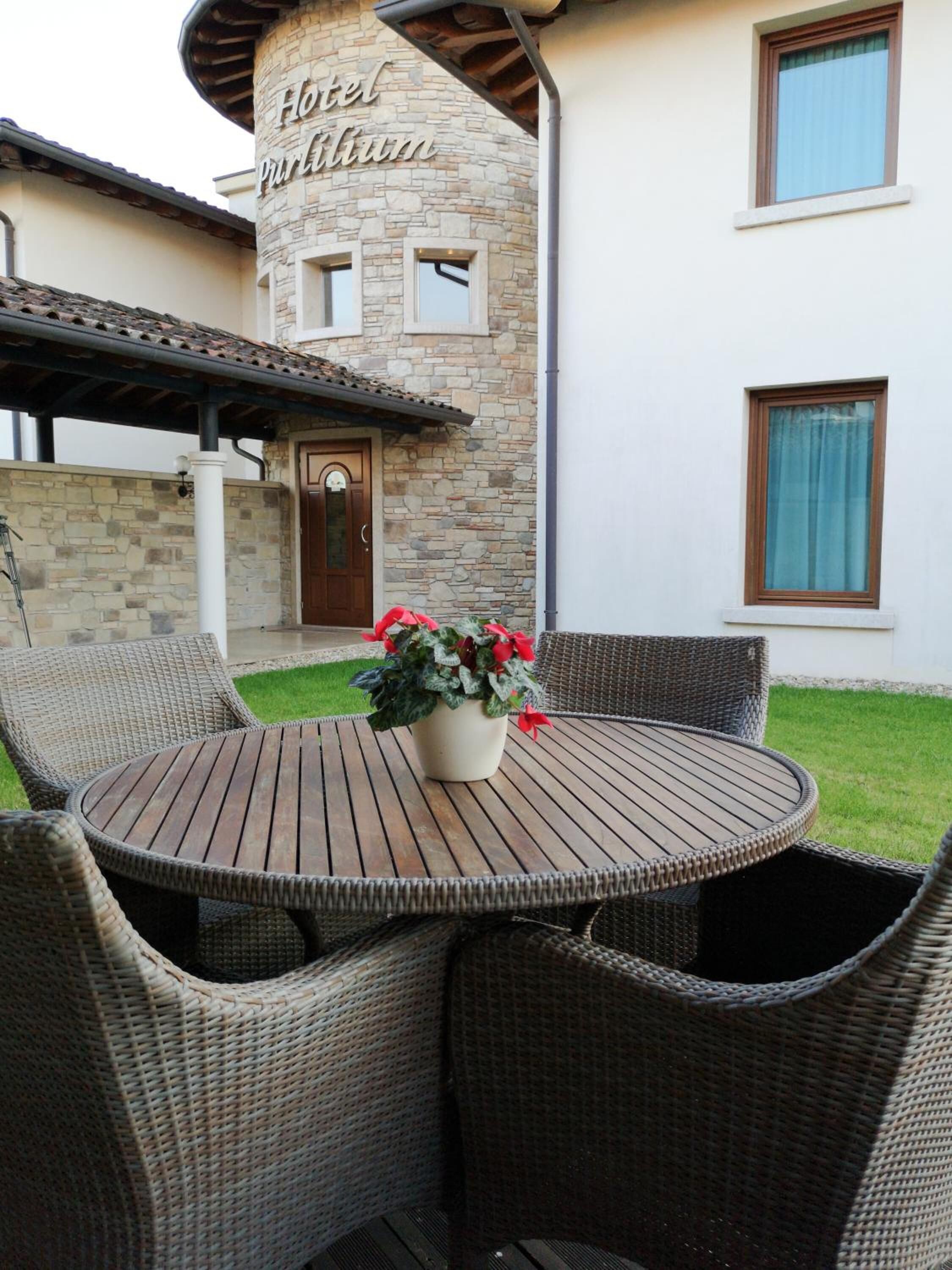 A round wooden table with wicker chairs and a potted plant with red flowers in front of a building with a stone facade and a sign that reads "Hotel Purlum".