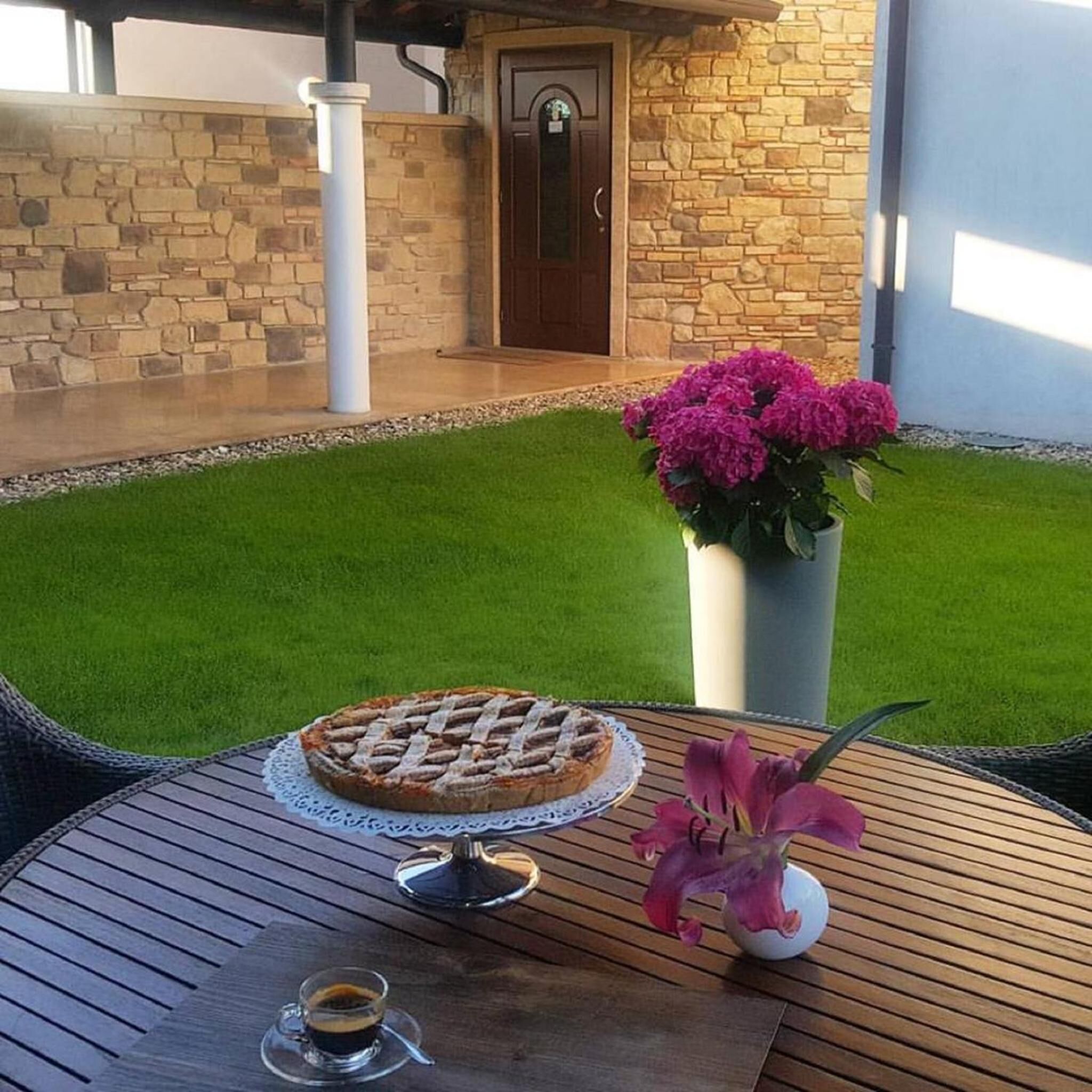 Un patio con una mesa redonda de madera, un pastel sobre un soporte, una taza de café y una planta en maceta con flores rosas.