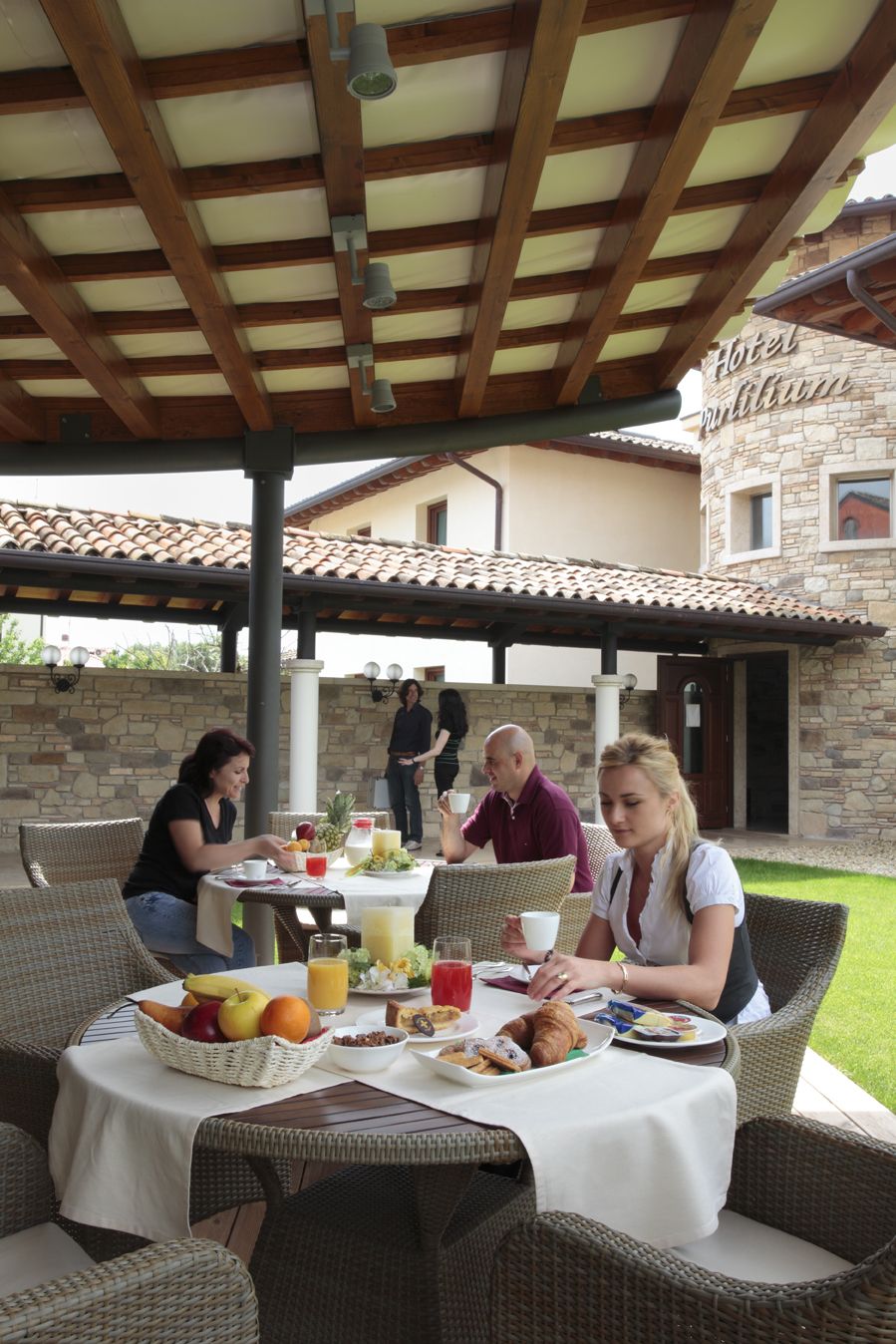 An outdoor dining area with wicker furniture, a stone building in the background, and a covered roof structure.