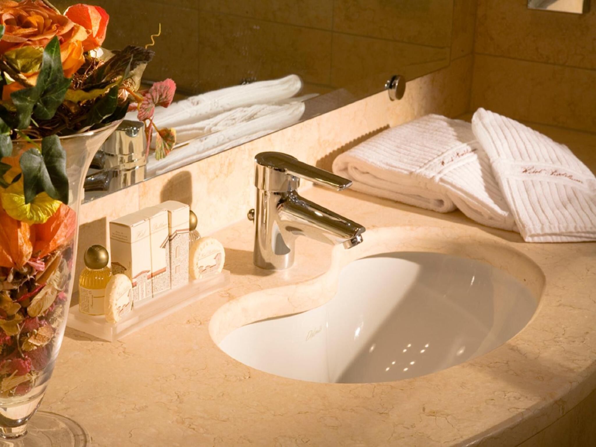 Un baño con lavamanos blanco, grifo cromado y espejo encima. Hay toallas y artículos de aseo en la encimera.