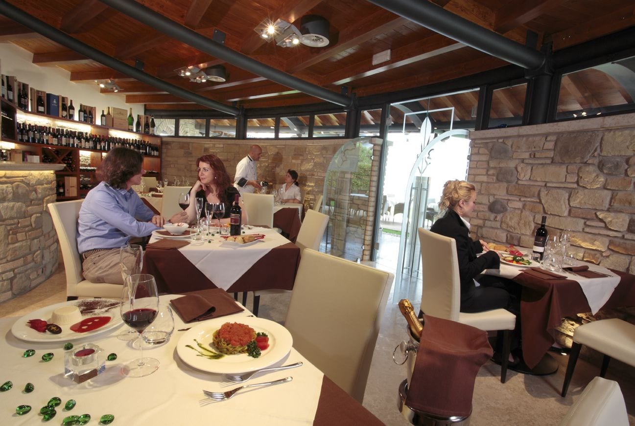 A restaurant with stone walls and wooden beams, tables set for dining, and a wine display in the background.