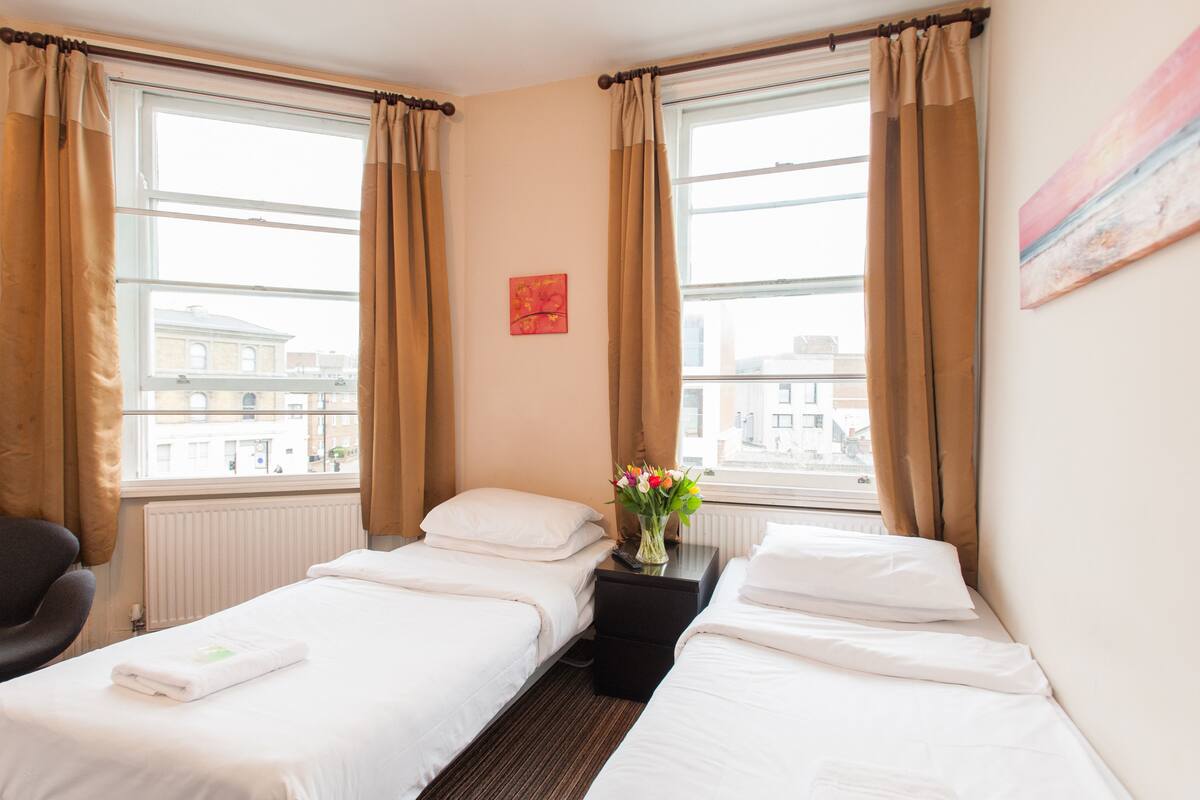 Ett hotellrum med två separata sängar, ett litet bord med en vas med blommor, en stol och en tavla på väggen.